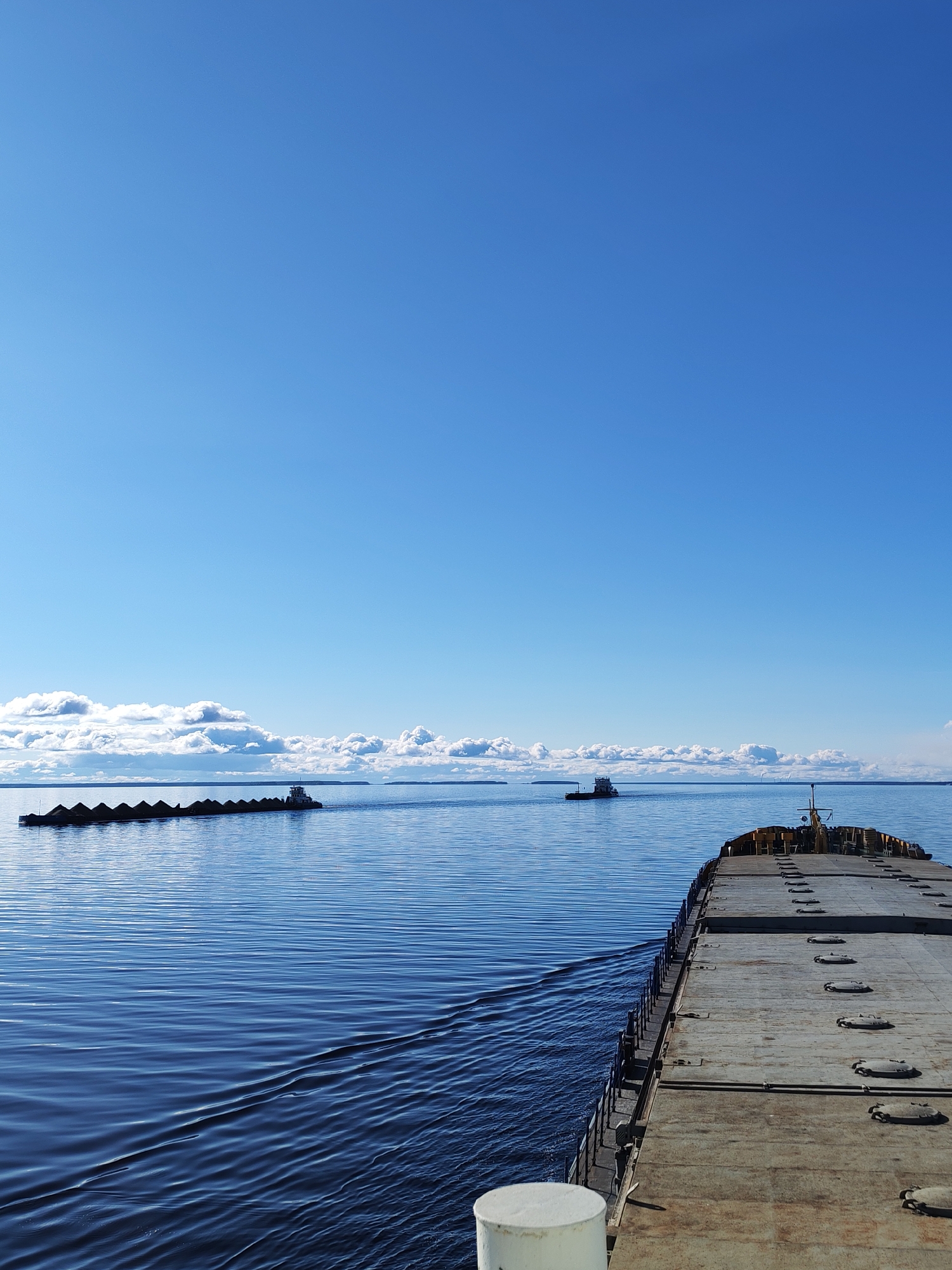 The image depicts a vast body of water under a clear blue sky with a few scattered clouds near the horizon. In the distance, two tugboats are visible: one towing a long barge laden with dark cargo, likely coal, and another smaller tugboat traveling alone. The scene is viewed from a barge, whose deck, with its manhole covers and metal railings, occupies the lower right corner of the frame. A white cylindrical object is visible in the immediate foreground, near the bottom edge. The water has gentle ripples and shows wake patterns from the vessels. The location is Mikhalevo, Russia.