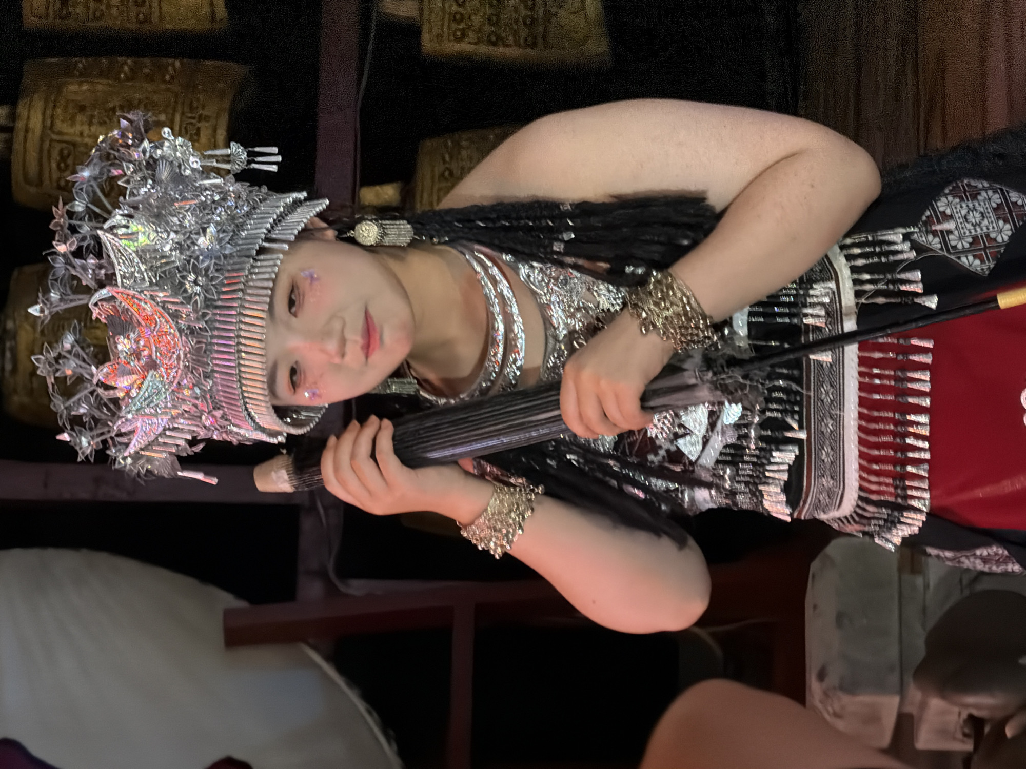 A young woman in traditional attire is holding a dark, cylindrical object with both hands, positioned in front of her. She is adorned with an elaborate silver headdress, multiple silver necklaces, ornate bracelets on both wrists, and dangling earrings. Her outfit consists of a black top with silver embellishments and a red skirt. She has glitter applied to her face. In the background, large, golden cylindrical objects are visible, appearing to be prayer wheels. The scene is dimly lit. The image was captured in Yuzhong District, China.