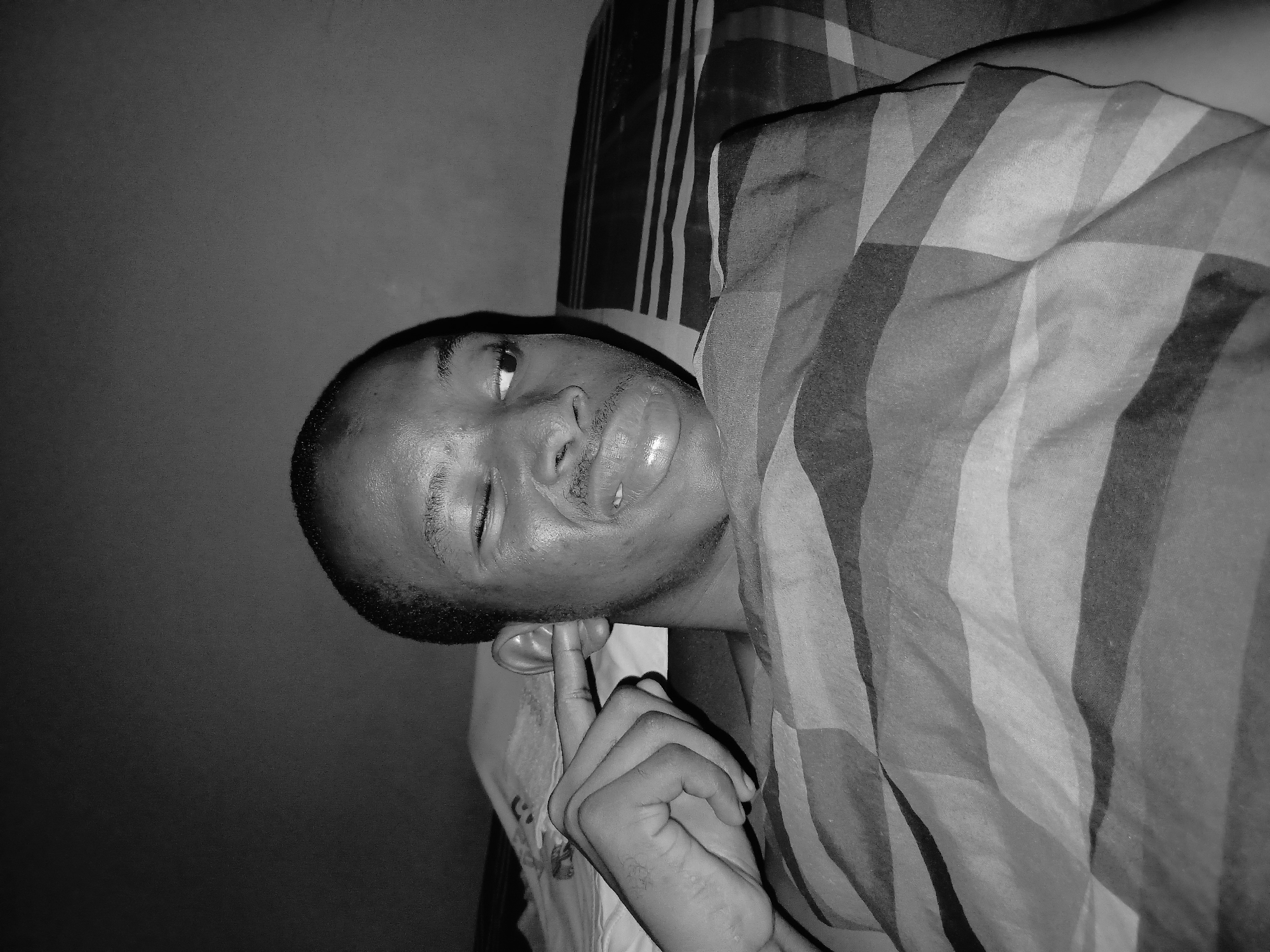 A black and white, medium close-up shot shows a young Black male lying in bed, his head turned to the right and his eyes partially closed as he winks. He is partially covered by a patterned blanket. His right hand is raised, with his index finger pointed towards his right ear, as if plugging it. His facial expression is one of playful mischief. The background appears to be a bedroom wall. The image does not contain any specific geographical markers to indicate its location in Lagos, Nigeria.