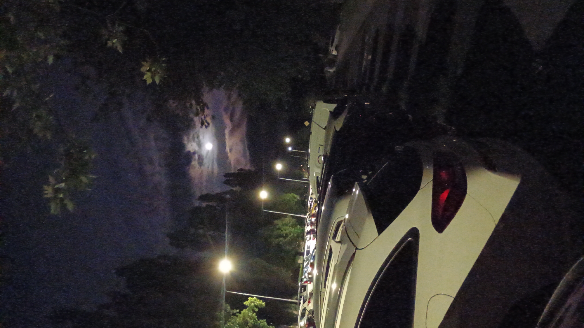 In Sokolov, Czechia, a nighttime scene unfolds in a parking lot illuminated by streetlights. The full moon shines brightly in the dark sky, partially obscured by clouds. Rows of cars are parked, with the foreground dominated by the rear of a white SUV featuring a glowing red taillight. Trees with dark foliage frame the scene, adding to the nocturnal ambiance.