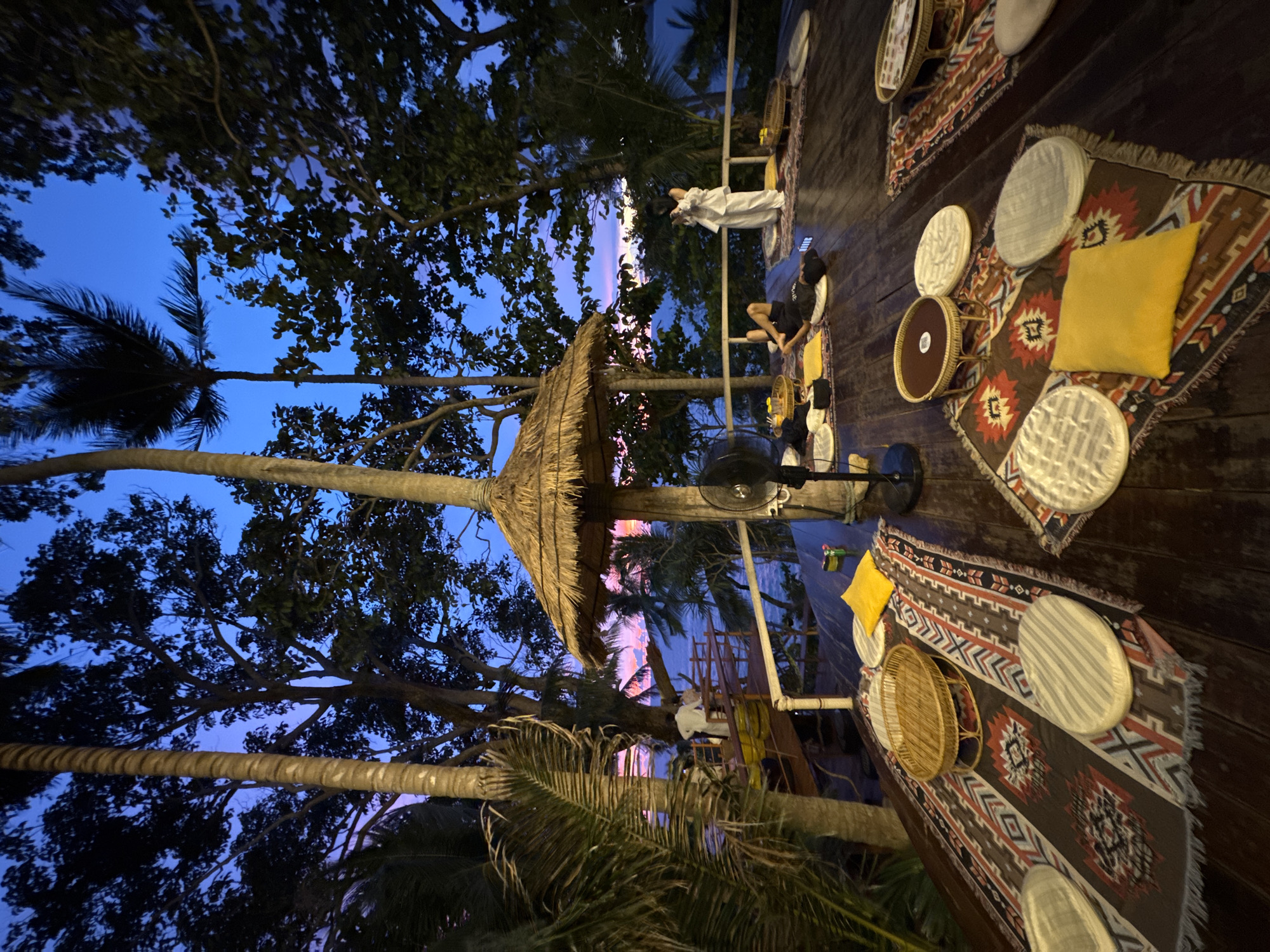 The image depicts an outdoor seating area at dusk, with a vibrant twilight sky transitioning from deep blue to shades of purple and pink. Large trees with dense foliage frame the scene, with palm trees and their fronds visible in the foreground, suggesting a tropical location. The seating area is on a wooden deck, adorned with colorful, patterned rugs. On the rugs are circular woven mats, some appearing to be placemats or bases for dishes. Several yellow cushions add pops of color to the seating arrangements.
In the background, two people are visible. One individual, dressed in white, stands near a railing. Another person, wearing dark clothing, is seated on a cushion. The setting appears to be a relaxed outdoor dining or lounging space, possibly a restaurant or bar, given the presence of a fan and what looks like a table or counter further back. The time of day is clearly indicated by the twilight sky. The overall mood is serene and tranquil, enhanced by the natural surroundings and the warm glow of the artificial lighting that is subtly illuminating the deck. The location context of Karon, Thailand, aligns with the tropical vegetation and the open-air, relaxed ambiance. There is no visible text in the image.