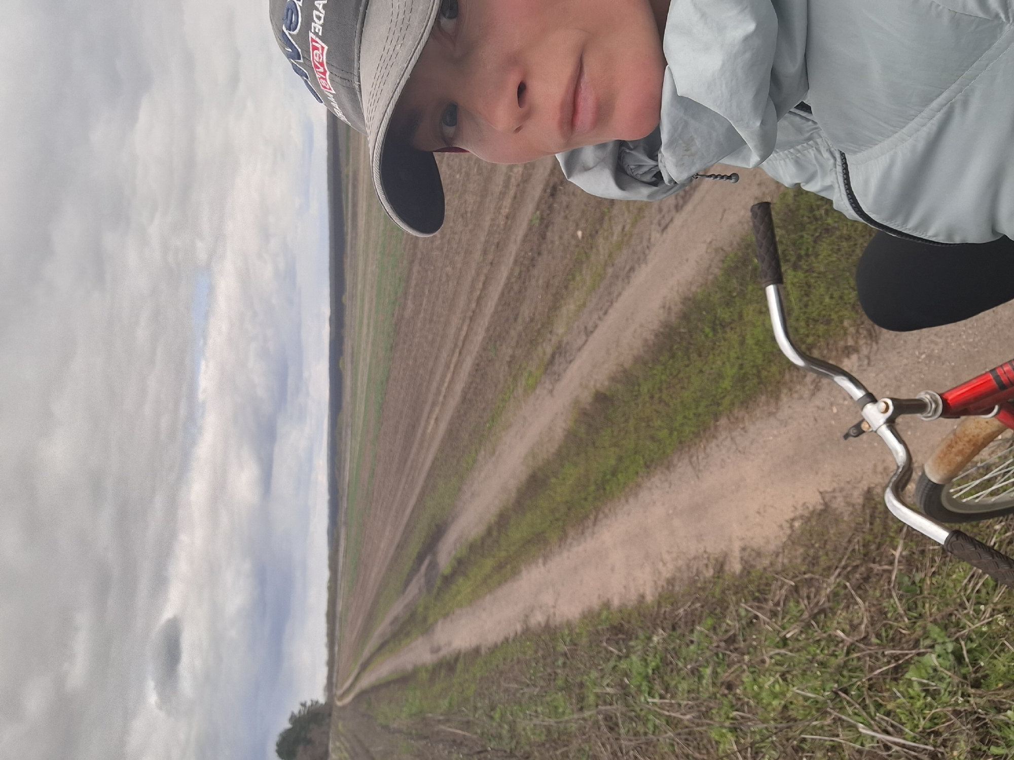 A selfie-style image captures a person on a bicycle in a vast rural landscape, identified as Treshchevka, Russia. The individual, seen from the chest up, wears a light blue or grey hooded jacket and a grey baseball cap. Their face is partially visible beneath the cap, with a neutral expression as they look directly at the camera. A red-outlined text or logo, possibly reading "...ADE RED...", is discernible on the side of the cap.
The person is positioned on a red bicycle, with its silver handlebars and black grips prominent in the lower and central foreground. The setting is dominated by expansive, flat agricultural fields featuring distinct furrows, suggesting tilled or recently planted land. A dirt path or track stretches into the distance, delineating the fields. Sparse green vegetation lines the path and is scattered within the fields. A faint line of trees marks the far horizon. The sky is overcast with varying shades of grey, indicating a cloudy daytime. The scene conveys a sense of serene travel through open countryside.