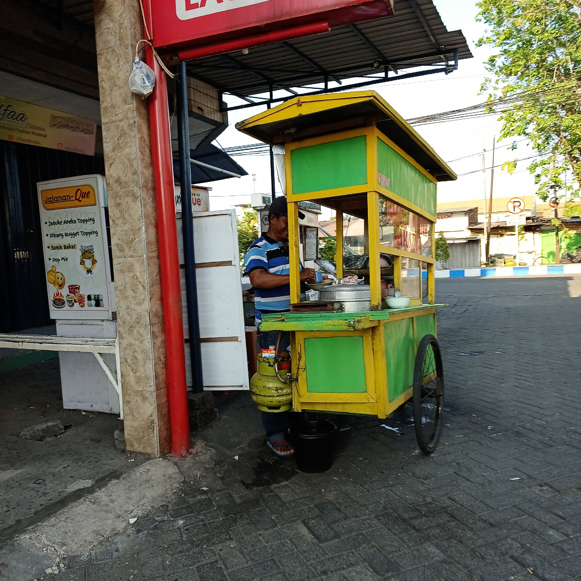 A street food vendor is preparing food at a colorful cart in Wedoro, Indonesia. The cart is yellow and green with a yellow roof. A man wearing a blue and white striped shirt and a dark cap is standing behind the cart, attending to a large pot on the stove. A green gas cylinder is attached to the cart. In front of the cart, a black bucket sits on the paved sidewalk. To the left of the cart, a sign displays a menu with items like "Jasuke Aneka Topping," "Pisang Nugget Topping," "Sosis Bakar," and "Pop Mie." The street is paved with interlocking bricks, and there are buildings and trees in the background. The scene appears to be on a street with a slight incline.