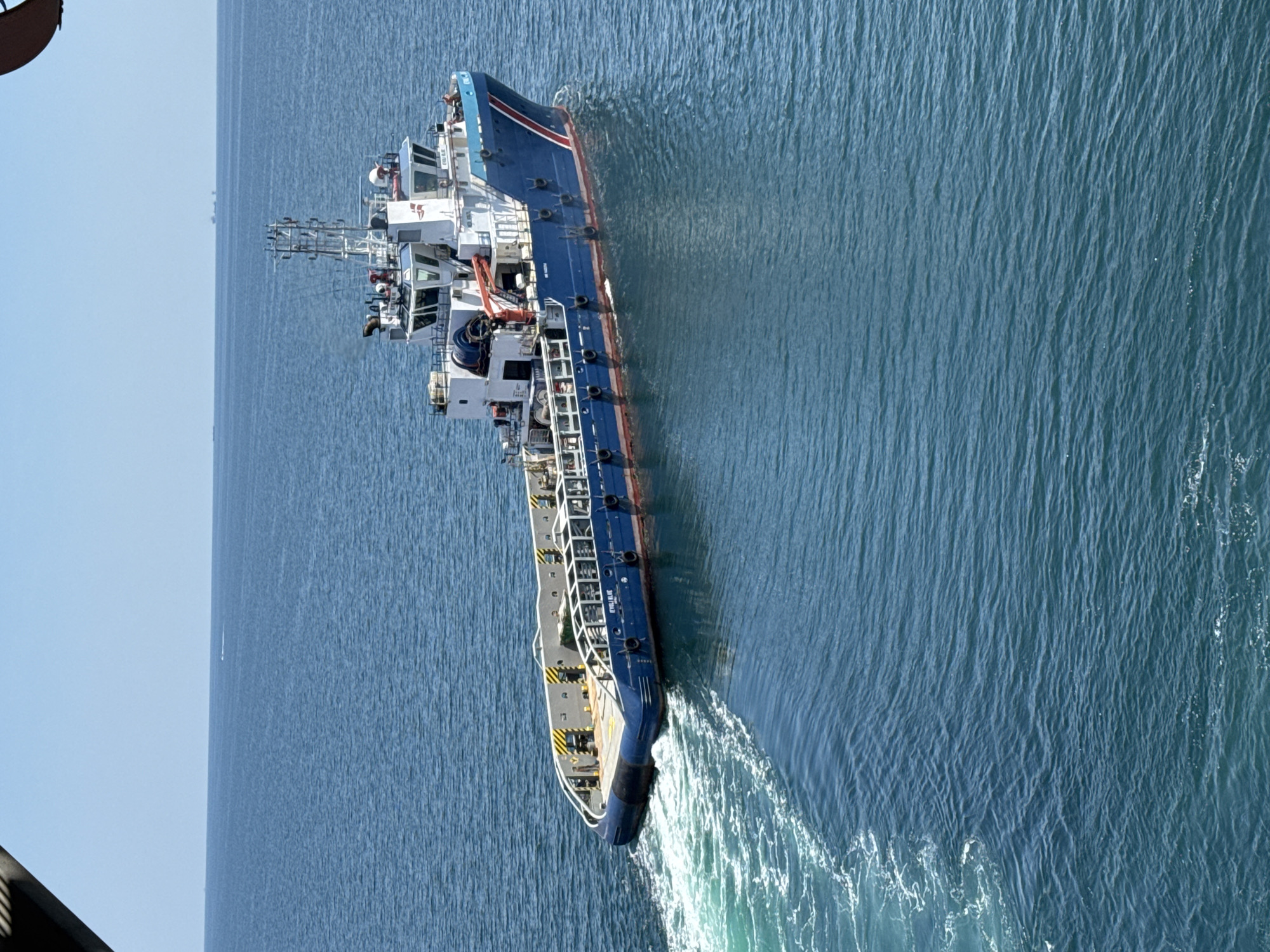 A blue and white vessel is seen from an elevated perspective, moving through calm blue water. The boat has a large deck area with yellow and black striped barriers and a crane. The bow of the boat is white with a blue stripe and a red trim line along the waterline. A wake is visible trailing from the stern of the boat. The horizon shows a clear blue sky with a few distant structures that appear to be oil rigs or other offshore installations. The image is taken from an aerial viewpoint, possibly from another boat or a helicopter.