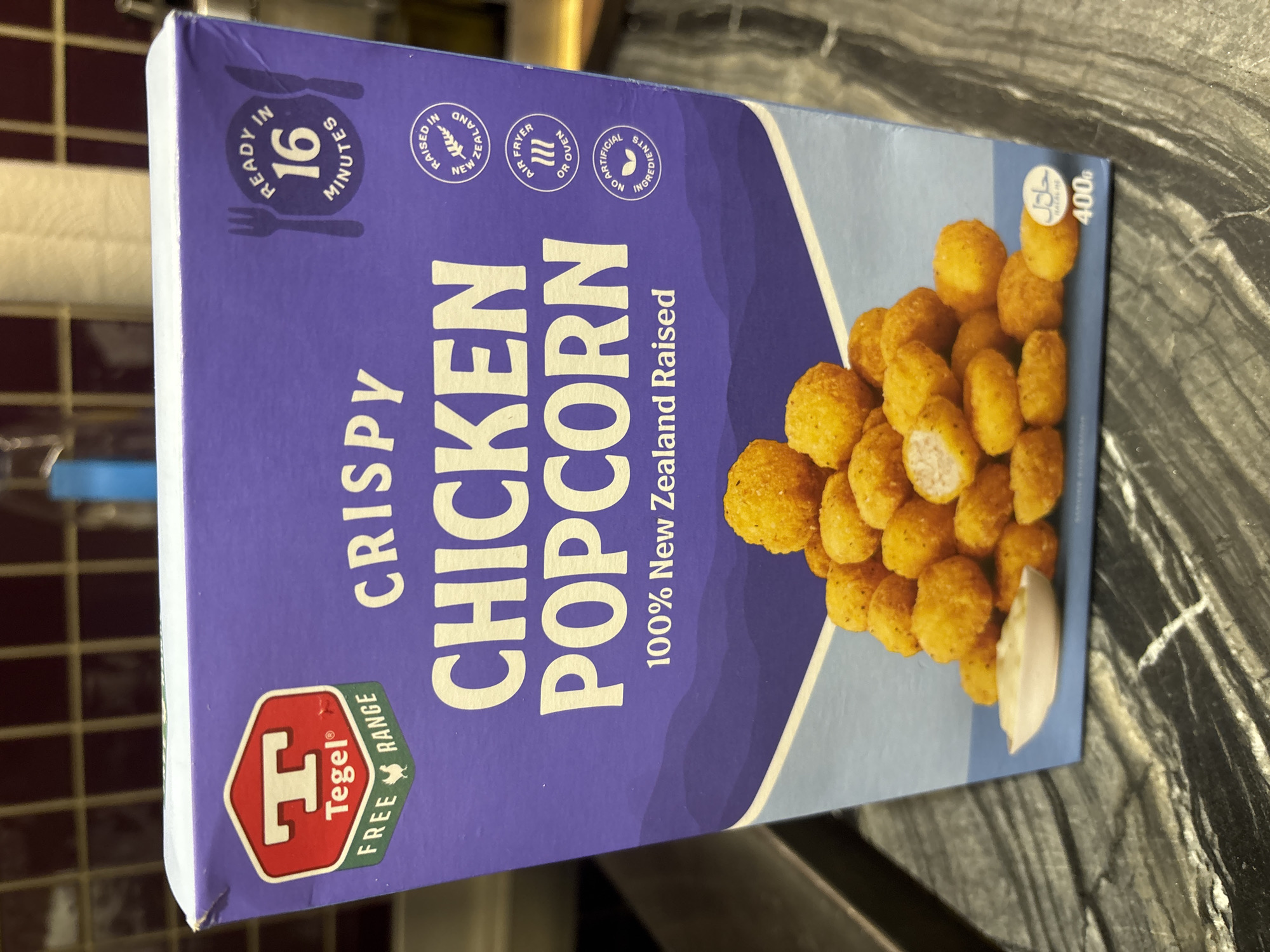 The image shows a close-up of a box of Tegel Crispy Chicken Popcorn. The box is predominantly purple with white text. The Tegel logo, featuring a red hexagon with a white "T" and the text "Tegel Free Range", is visible on the top left. Text on the box reads "CRISPY CHICKEN POPCORN 100% New Zealand Raised". To the right of the text are circular icons indicating "READY IN 16 MINUTES", "RAISED IN NEW ZEALAND", "AIR FRYER OR OVEN", and "NO ARTIFICIAL INGREDIENTS". The bottom of the box displays an image of chicken popcorn pieces with a small dish of sauce. A "HALAL" certification symbol and "400g" are visible on the bottom right. The background shows a tiled wall and a dark countertop. There is no indication of the city of Dubai, United Arab Emirates, in the image.