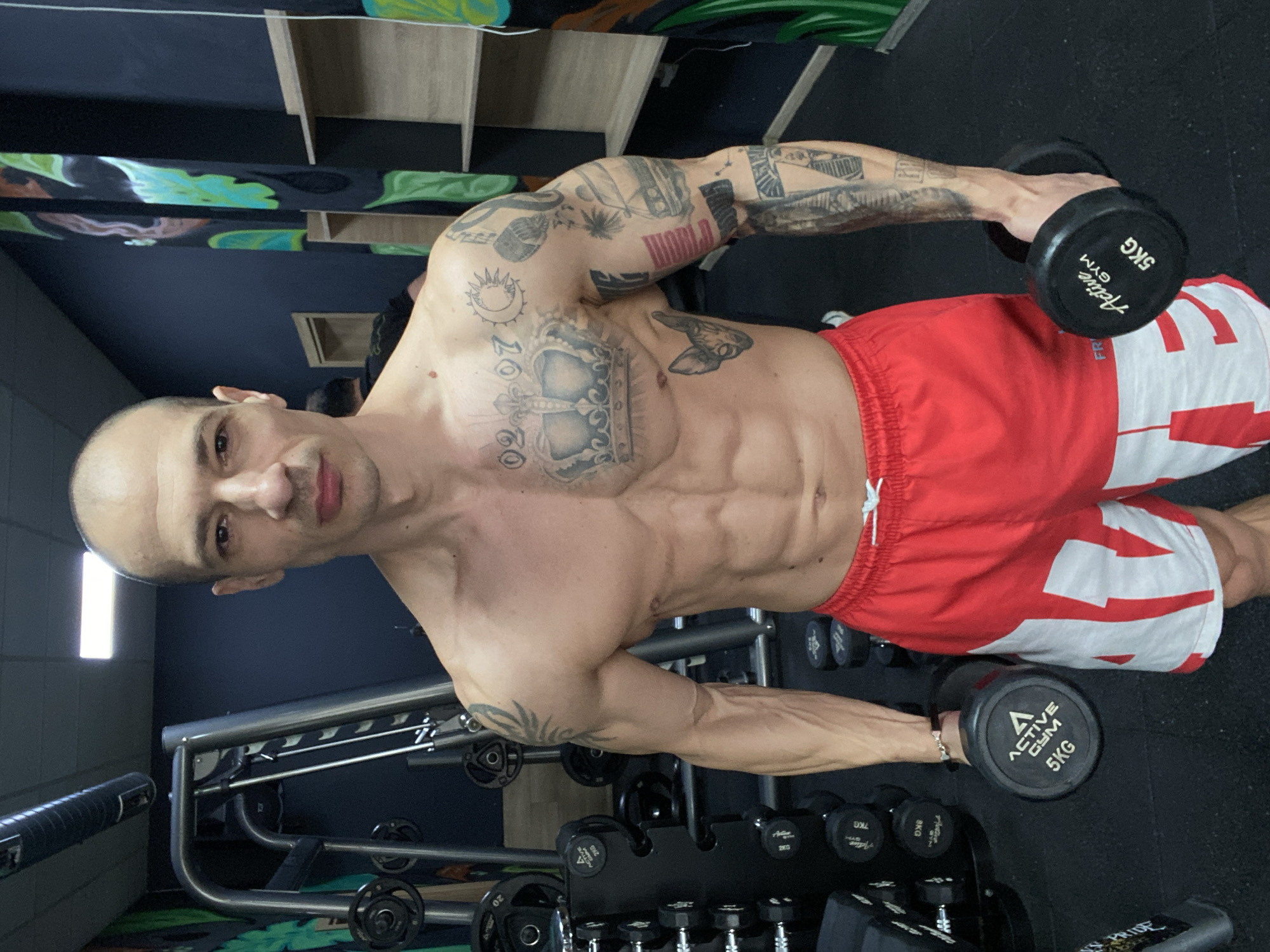A muscular man is posing while holding two 5kg dumbbells in a gym. He is shirtless, wearing red and white shorts. The man has tattoos on his arms and chest. The gym contains a rack of dumbbells and other gym equipment. The location may be in Pleven, Bulgaria.
