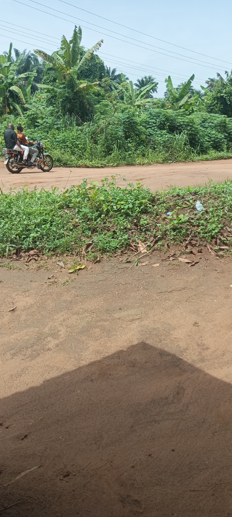 The scene is an outdoor view in Amauzu, Nigeria. A motorcycle with two occupants is moving along a dirt road. The driver is wearing a printed shirt and white pants. The passenger is wearing a dark shirt and is positioned behind the driver. The road is bordered by dense green vegetation including banana trees. A portion of the foreground is blurred. Electrical cables can be seen crossing above the road. The lighting suggests a bright, sunny day.