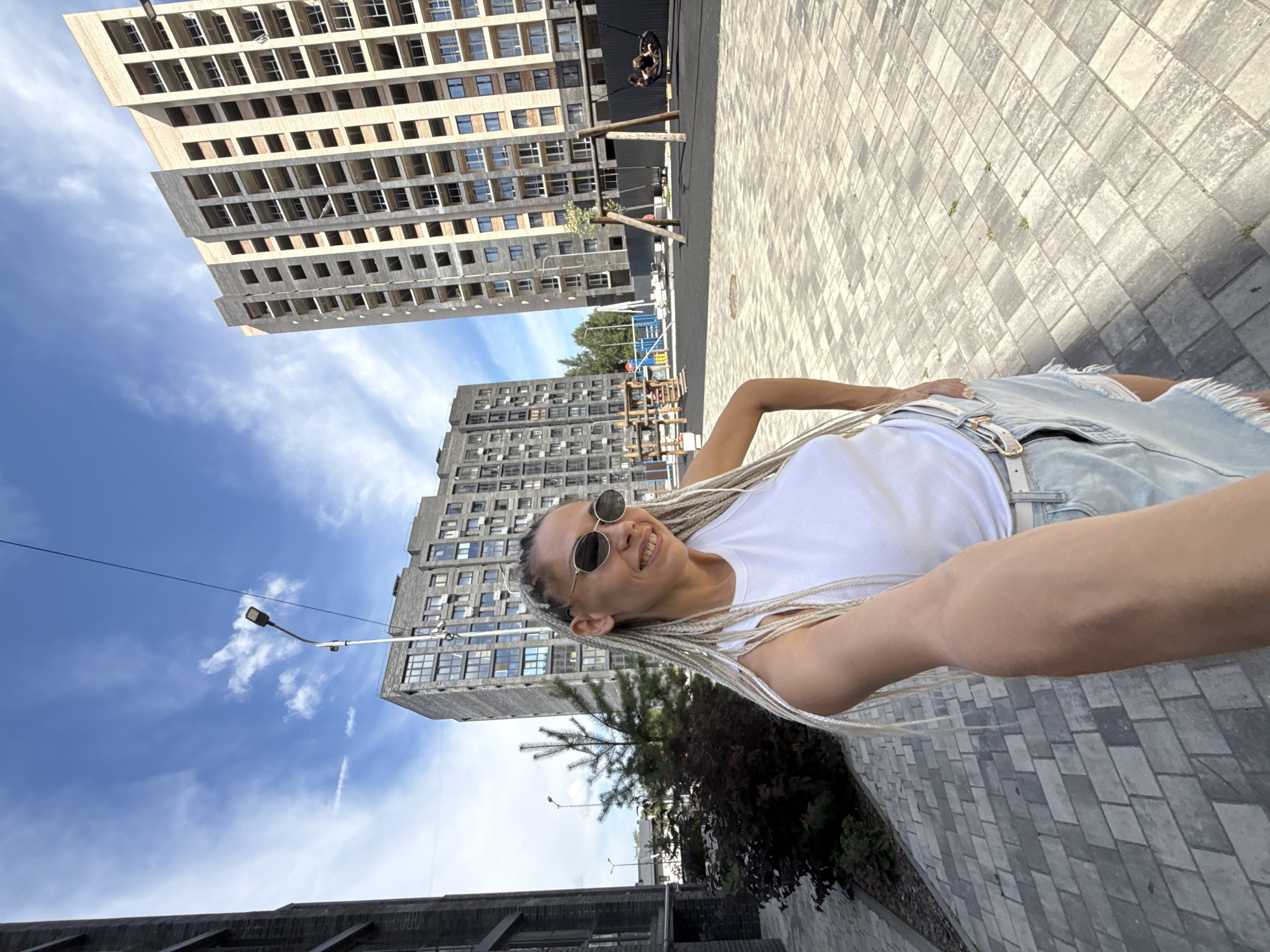 A selfie taken on a paved courtyard in a residential area of Rusanovo, Russia shows a woman with blonde braided hair, wearing a white tank top, denim skirt, and sunglasses. She is smiling and holding the camera at arm's length. In the background, there are two multi-story apartment buildings under construction, a playground with swings, and a street lamp. The sky is blue with scattered white clouds.