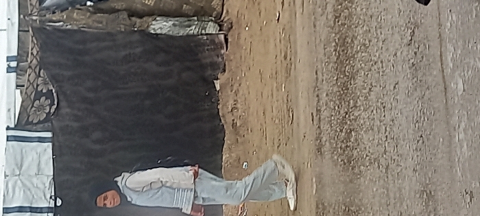 In Khan Yunis, Palestinian Territory, a young person walks left-to-right across an unpaved, dirt and gravel path. The individual is dressed in a black hijab, a light-colored long-sleeved top, a light blue sleeveless vest, light-colored trousers, and white athletic shoes, maintaining a slight smile and looking towards the viewer. The background features several makeshift structures constructed from draped fabrics and tarpaulins, including a dark grey section, a white tarp with two vertical dark stripes, and a patterned fabric displaying dark floral designs. Scattered debris is visible on the ground behind the person.