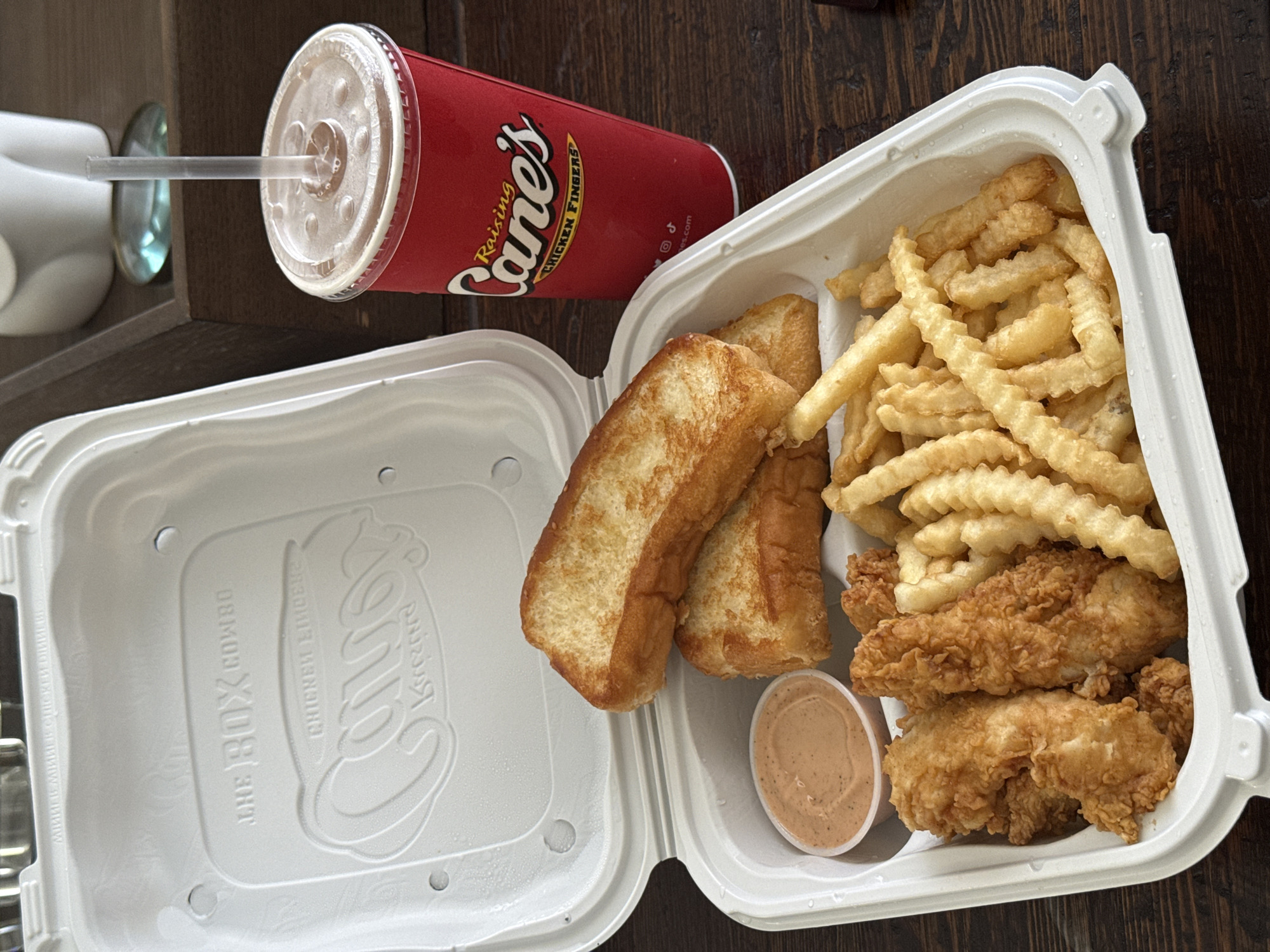 On a dark brown wooden surface, a "Raising Cane's" meal is displayed, potentially in Chula Vista, California, judging by the brand's locations. The meal consists of an open white styrofoam container containing crinkle-cut french fries, three fried chicken strips, two pieces of buttered toast, and a small container of sauce. Next to the container is a red "Raising Cane's" drink cup with a clear lid and a straw. A white object with a clear disc is vaguely visible in the background.