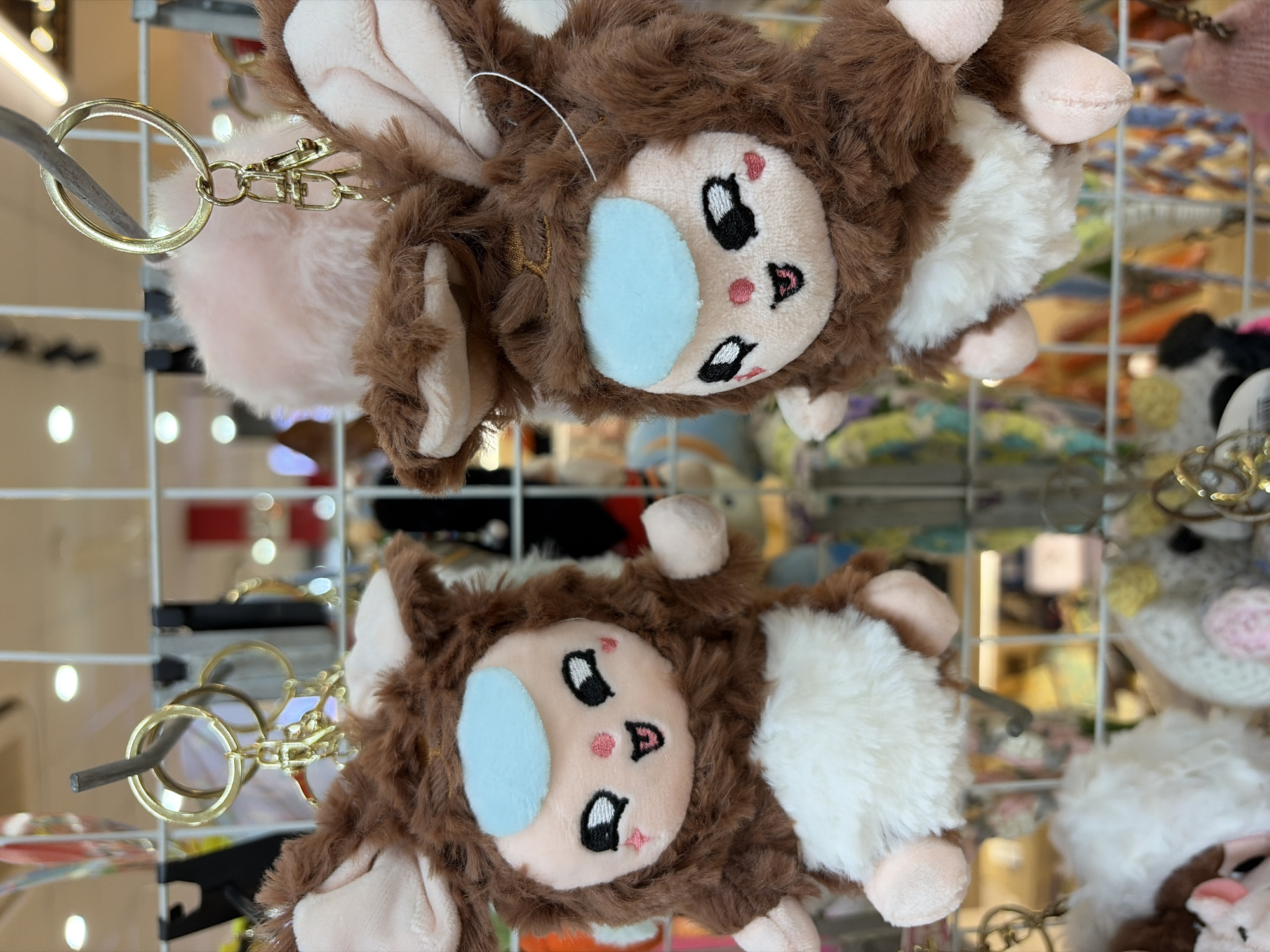 The image displays two plush toys with brown fur and pink ears, resembling small creatures, hanging from a metal grid. Both toys have a light blue patch on their forehead, round black eyes with white highlights, pink cheeks with rosy blush marks, and open mouths with visible pink tongues and white teeth. They also have white fluffy bellies and pink paws. Both toys are attached to gold-colored keychains with clips and rings. The background consists of a blurred retail environment with shelves, other merchandise, and lighting. The image is a medium shot focused on the two plush toys.