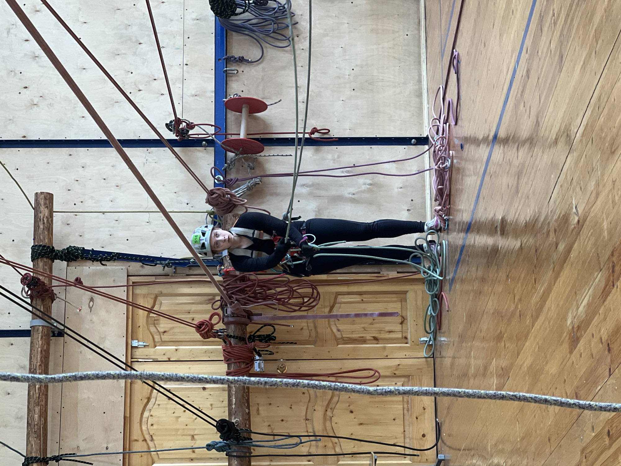 A woman in a climbing harness and helmet is suspended by ropes in an indoor rope course. The course consists of wooden posts, ropes of various colors, and wooden platforms. The environment appears to be a gymnasium or indoor recreation facility with wooden walls and floors. There are doors in the background and metal safety clips hanging from the ropes. The location is Chaikovsky, Russia.