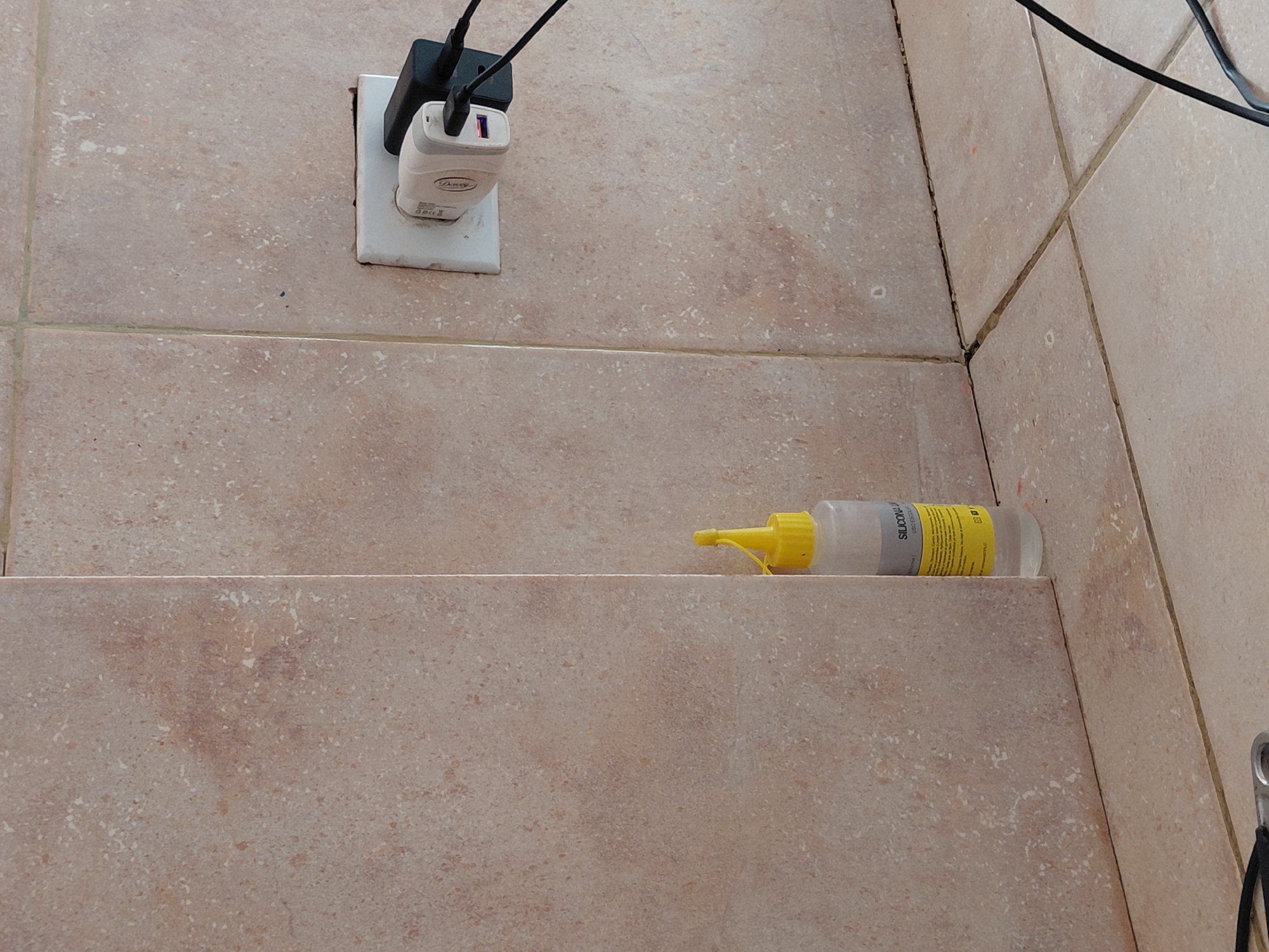 The image captures an indoor scene, likely in Santa Ana, Venezuela, featuring a tiled wall with an electrical outlet and a bottle resting on a lower tiled surface.
In the upper left, a white electrical outlet is mounted on the light beige-colored tiled wall. Two chargers are plugged into the outlet: a black rectangular charger at the top and a smaller white charger below it. Black cables extend upwards and to the right from both chargers. The white charger has the brand name "Provy" and some small technical specifications, including "5.0V" and electrical symbols, printed on its side. The tiles are a speckled, light brownish-beige color with visible grout lines.
Towards the bottom right, a clear plastic bottle with a bright yellow pointed cap and nozzle is resting horizontally on a step or shelf made of similar light beige tiles. The bottle has a yellow label wrapped around it, on which the text "SILICONA LIQUIDA" is clearly visible, indicating it is liquid silicone. Smaller lines of text, likely product instructions or details, are also present on the label, along with what appears to be a small house symbol.
The tiles throughout the scene exhibit a slightly textured appearance with varying shades of beige and brown speckles. Some of the grout lines, particularly in the corner where the wall meets the adjacent tiled surface, show signs of discoloration or dirt. No human activity is depicted, but the plugged-in chargers suggest electronic devices are being powered or charged. The overall impression is that of a utility or service area, possibly a bathroom or kitchen, given the tiled surfaces and the presence of silicone sealant.