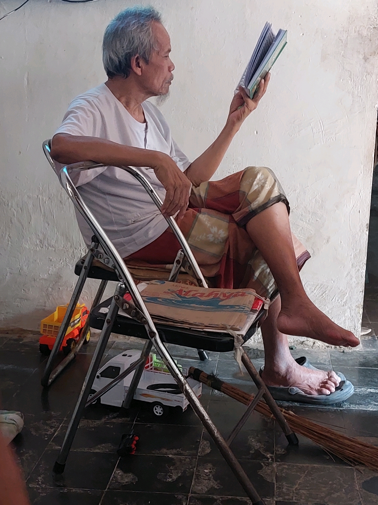 A man with graying hair and a goatee sits on a metal folding chair reading a book. He is wearing a white t-shirt and a patterned sarong. His right leg is crossed over his left, and his right foot is resting on a grey sandal. A broom is placed on the floor next to the chair. A stack of magazines or papers is on the seat of the chair. In the background, a white toy truck and a yellow and orange toy truck are on the floor. The man appears to be indoors, with a white wall behind him. The scene is likely in Sarangan, Indonesia, given the provided location context, suggesting a warm climate and possibly a home or communal area.