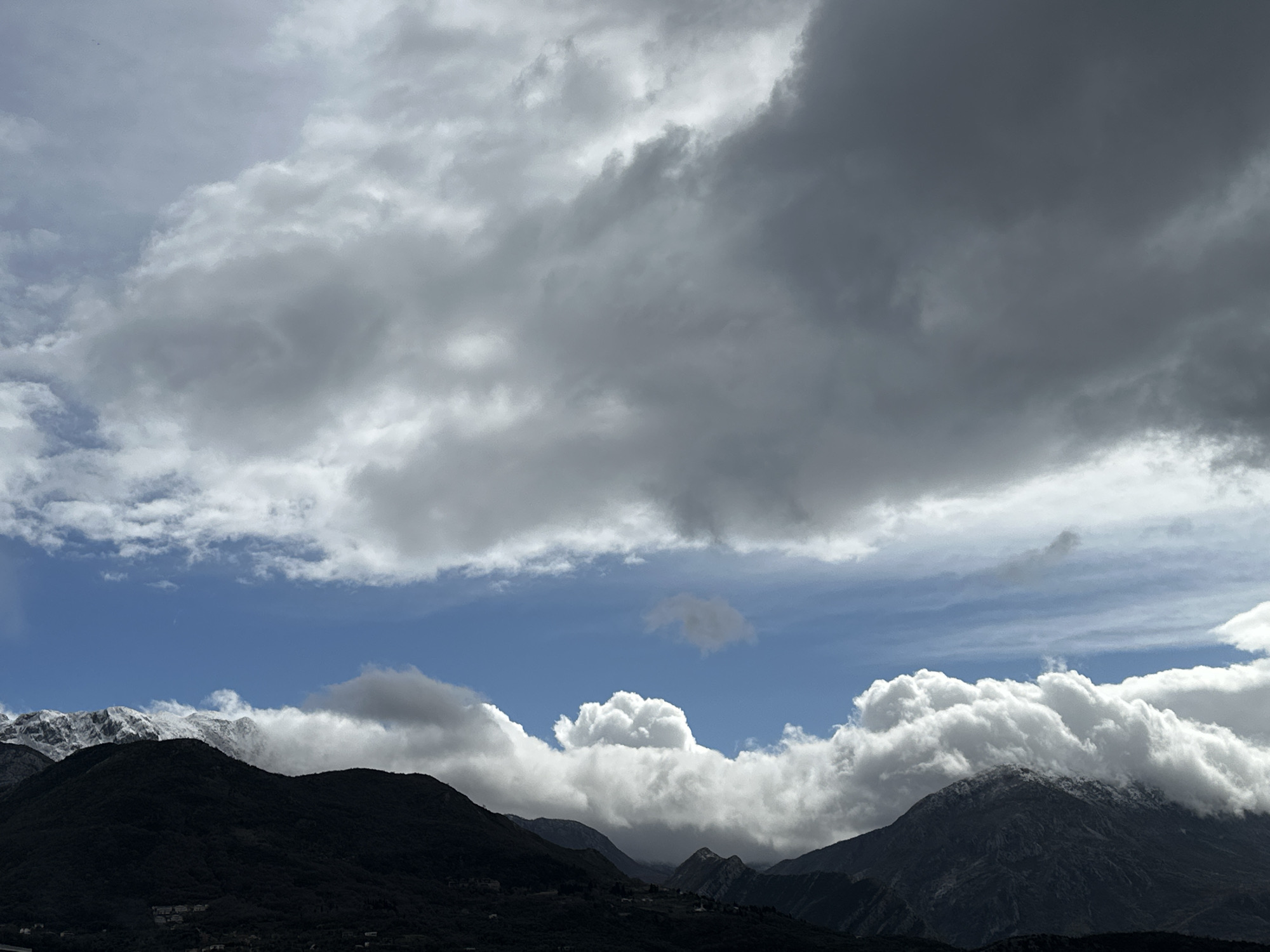 The image depicts a panoramic view of mountainous terrain under a dynamic sky, consistent with a perspective from or within the vicinity of Šušanj, Montenegro. The lower half of the frame features dark, tree-covered mountain slopes, with scattered light-colored residential structures faintly visible on their lower elevations. The upper peaks of the mountains in the background are partially covered with snow. The sky occupies the upper two-thirds of the image, characterized by a distinct horizontal band of clear blue sky in the middle, flanked by extensive, dense grey cloud formations above and clusters of bright, white cumulus clouds along the mountain ridges and within the mid-sky. No individuals, overt actions, or interactions are present in the scene.