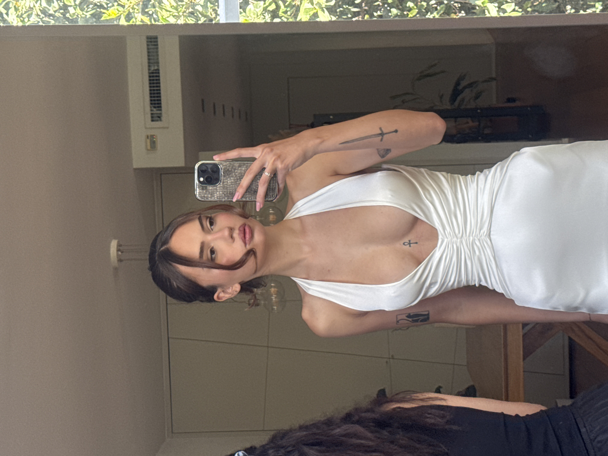 A young woman is captured in a mirror selfie in an indoor setting, likely in Tel-Aviv, Israel. The image is taken from the chest up, with the woman posing slightly angled. She is wearing a form-fitting white dress with a plunging neckline and ruched detailing on the torso. Her dark hair is styled, with some strands framing her face, and she looks directly at the camera with a neutral expression.
She holds a smartphone with a distinctive textured, possibly patterned, case in her right hand to take the photograph. Visible tattoos include a sword and a geometric shape on her upper right arm, an ankh symbol on her chest, and another framed tattoo on her left forearm.
The mirror's reflection shows a bright daytime environment, with a window at the top revealing lush green foliage outdoors, suggesting natural light fills the room. Other reflections include an air ventilation grate on the wall and various indistinct indoor elements, hinting at a modern living space or hotel room. A portion of another person's dark, curly hair and shoulder is visible in the lower part of the reflection.