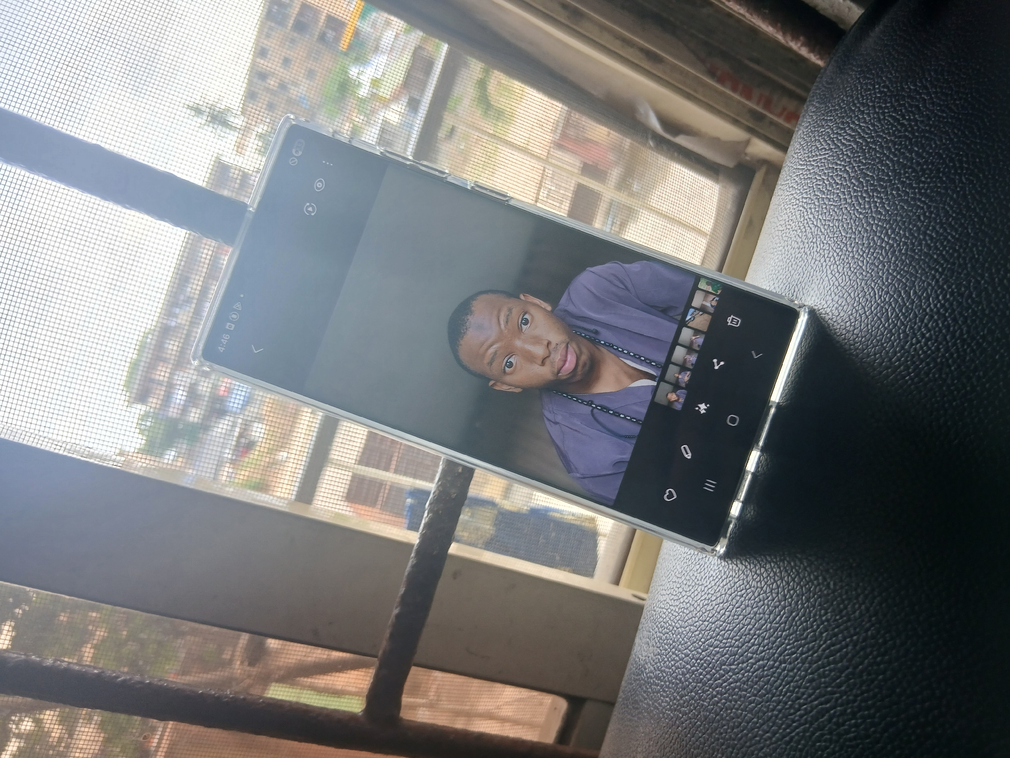 A phone is laying on a black surface. The phone's screen is displaying a photo of a man wearing a purple shirt and beaded necklace. The background of the image includes a window with a screen, showing a cityscape that is potentially in Lagos, Nigeria. The cityscape includes buildings and greenery.
