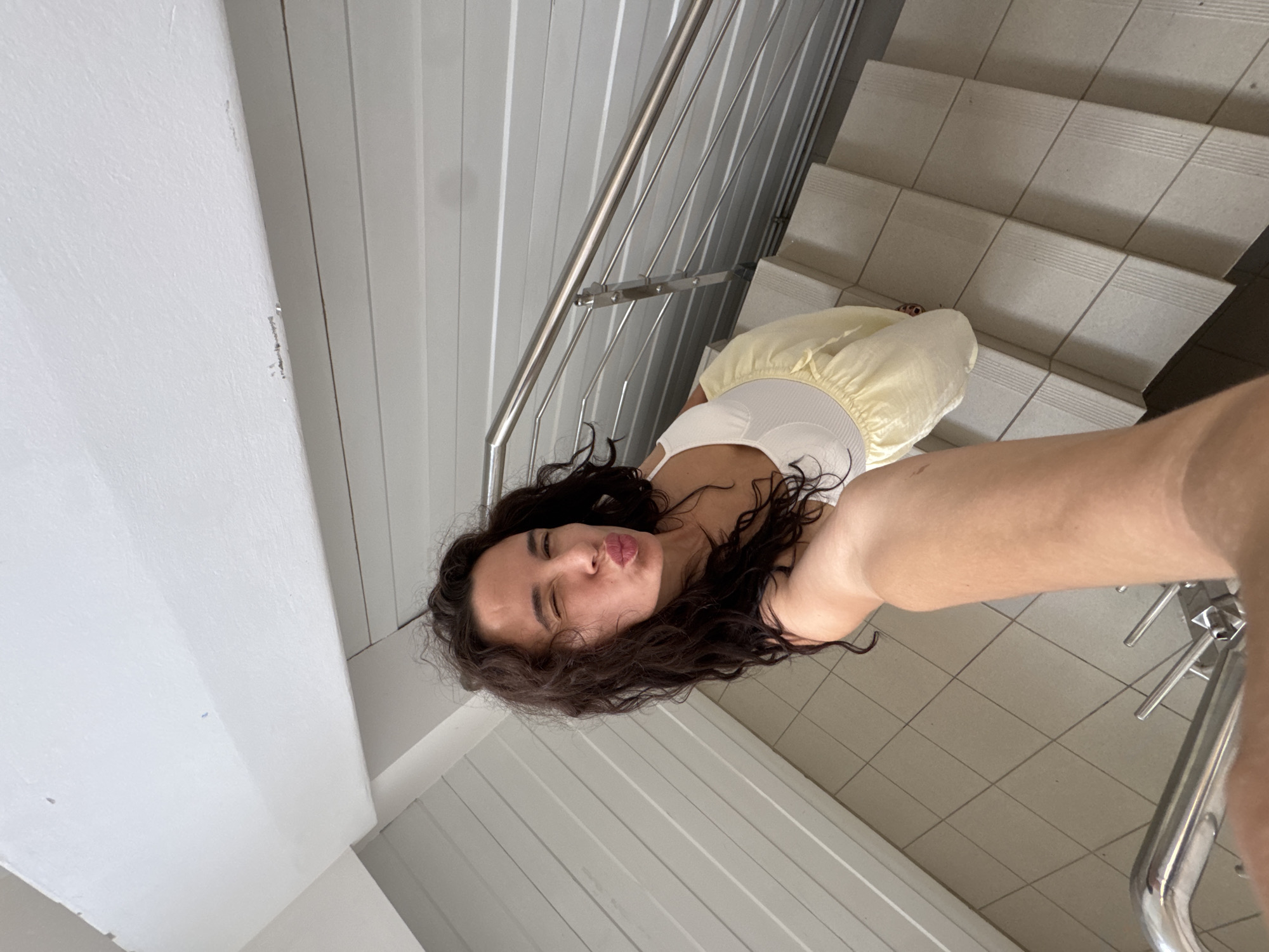 A selfie is taken from a first-person perspective on a staircase. The subject, a woman with long, dark, curly hair, is looking at the camera and puckering her lips, winking with her left eye. She is wearing a white top and a pale yellow skirt. The staircase has light-colored tiled steps and a metal railing. The walls on either side of the staircase are paneled with white horizontal planks. The image does not provide any specific visual cues to identify its location within the city of Chaikovsky, Russia.