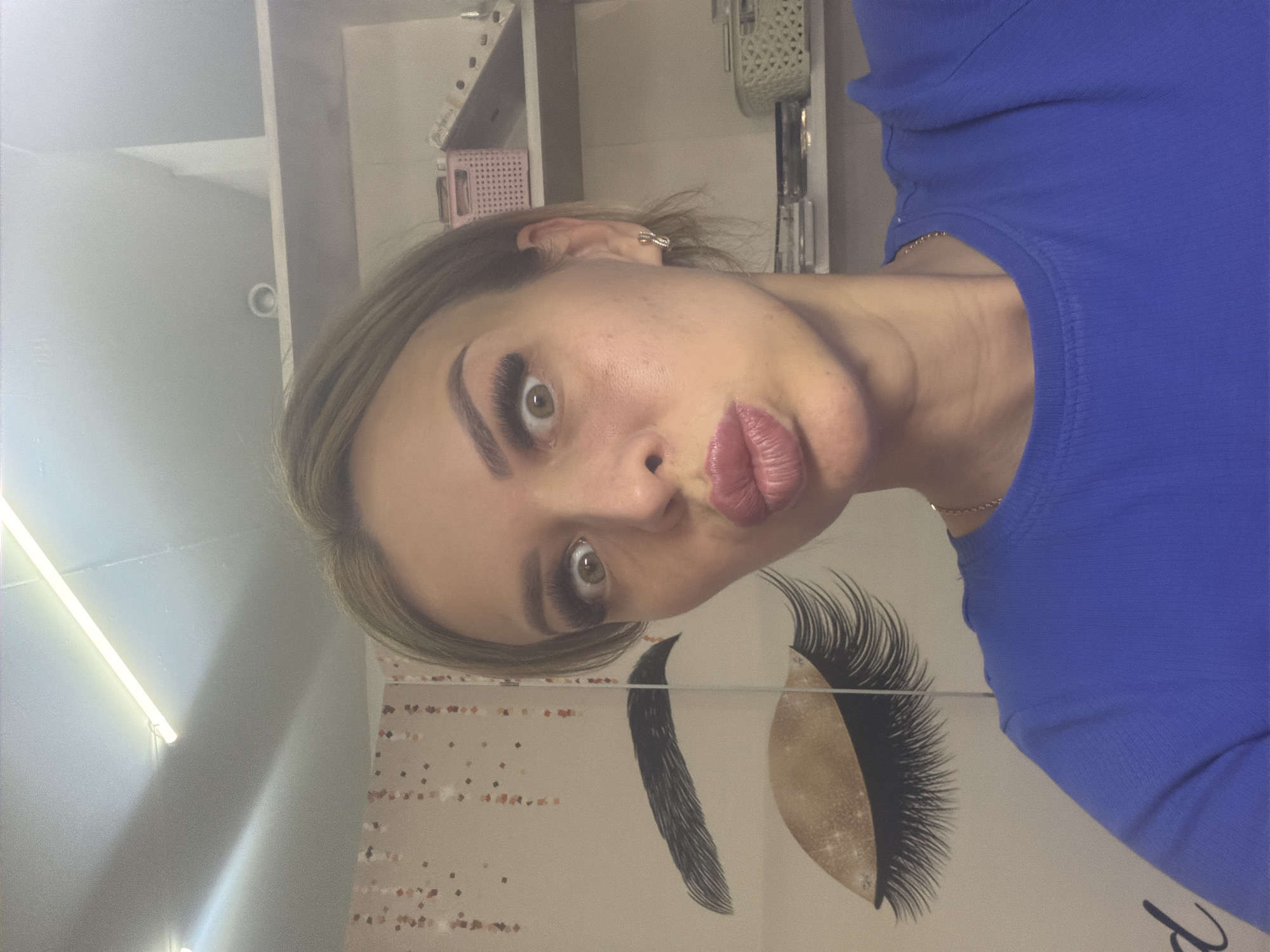 A woman with blonde hair and makeup is taking a selfie in what appears to be a beauty salon or similar establishment. She is making a kissing face with her lips puckered and her eyes wide open. She is wearing a blue shirt and a gold necklace. In the background, there is a wall with a graphic of eyelashes and a shelf with various beauty products. The lighting is bright, suggesting artificial light sources like fluorescent tubes. There is no indication of the specific location within Kyiv, Ukraine.