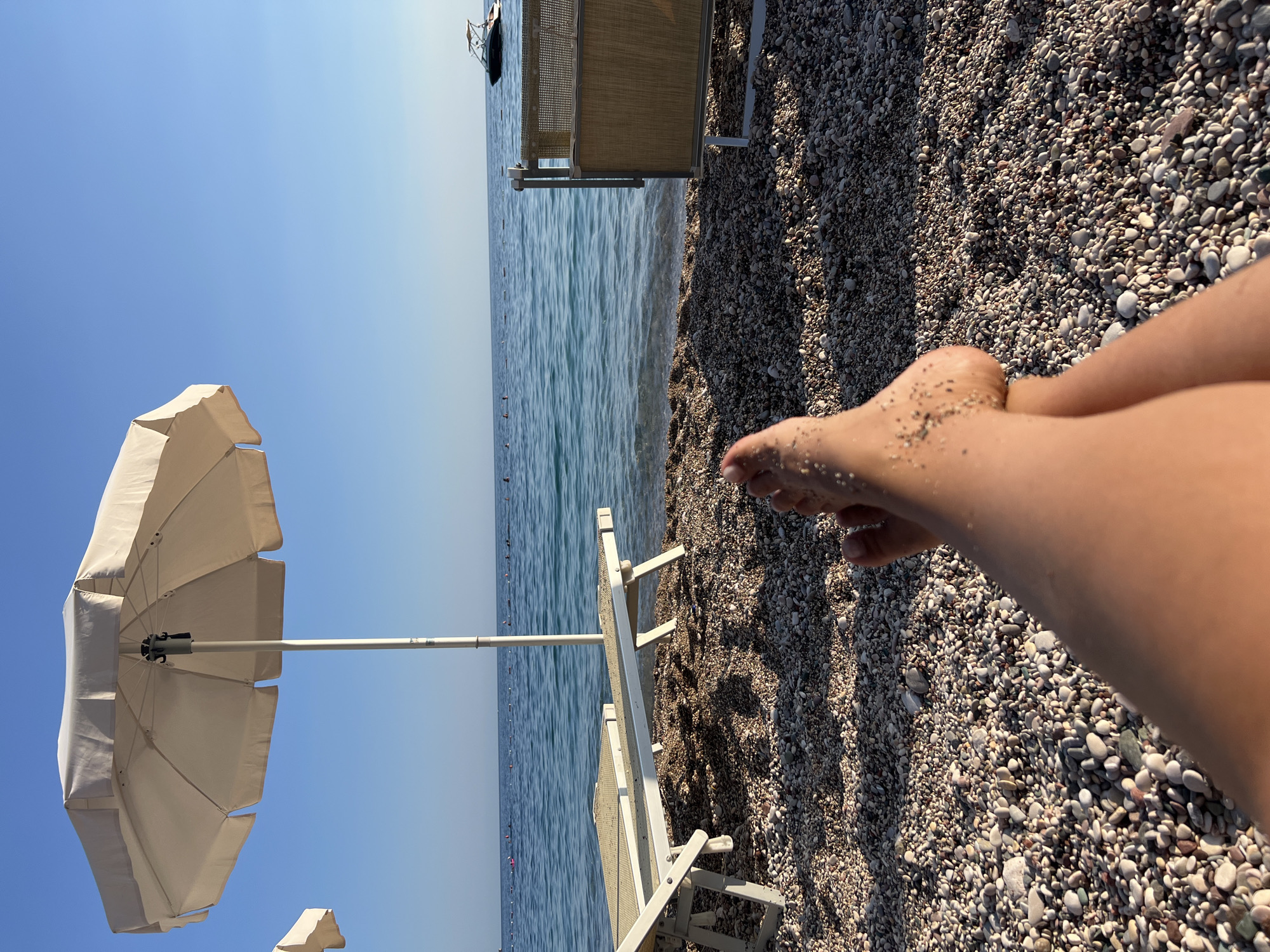 The media file depicts a person's legs and feet resting on a pebble beach, with a beach umbrella and lounge chairs visible in the background. The ocean stretches out to the horizon under a clear blue sky, suggesting a sunny day. The sand on the person's feet indicates they have been enjoying the beach. The scene evokes a sense of relaxation and leisure. The time of day appears to be daytime, given the bright sunlight casting shadows on the pebbles. The location is a beach in Kamenovo, Montenegro, as indicated by the context. There is no visible text in the image.