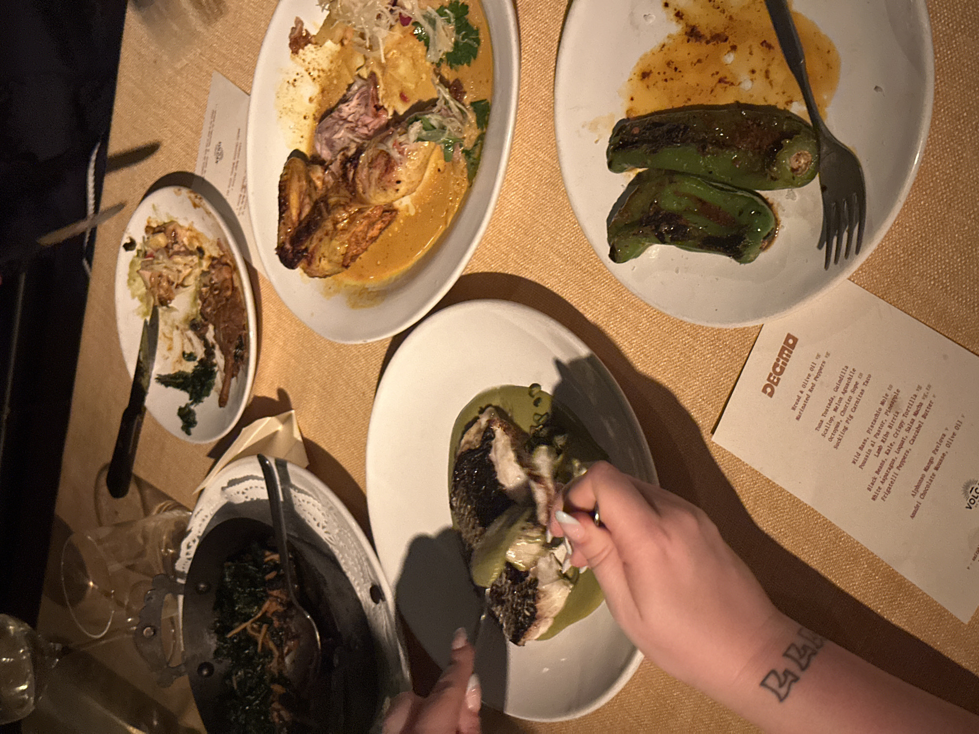 The image depicts a meal at Decimo restaurant in London, United Kingdom, based on the restaurant's menu visible in the shot. The view is from above, capturing various plates of food arranged on a table covered with a neutral-colored surface that has a subtle texture. Plates contain different dishes, including what appears to be fish with a green sauce, meat with a yellow sauce, blistered peppers, and another dish with various ingredients. Cutlery is visible near the plates, as well as a wine glass. Part of a person's forearm with a tattoo is visible, and hands are shown manipulating cutlery, suggesting someone is eating or preparing to eat. The lighting in the scene appears dim, creating a relaxed atmosphere.