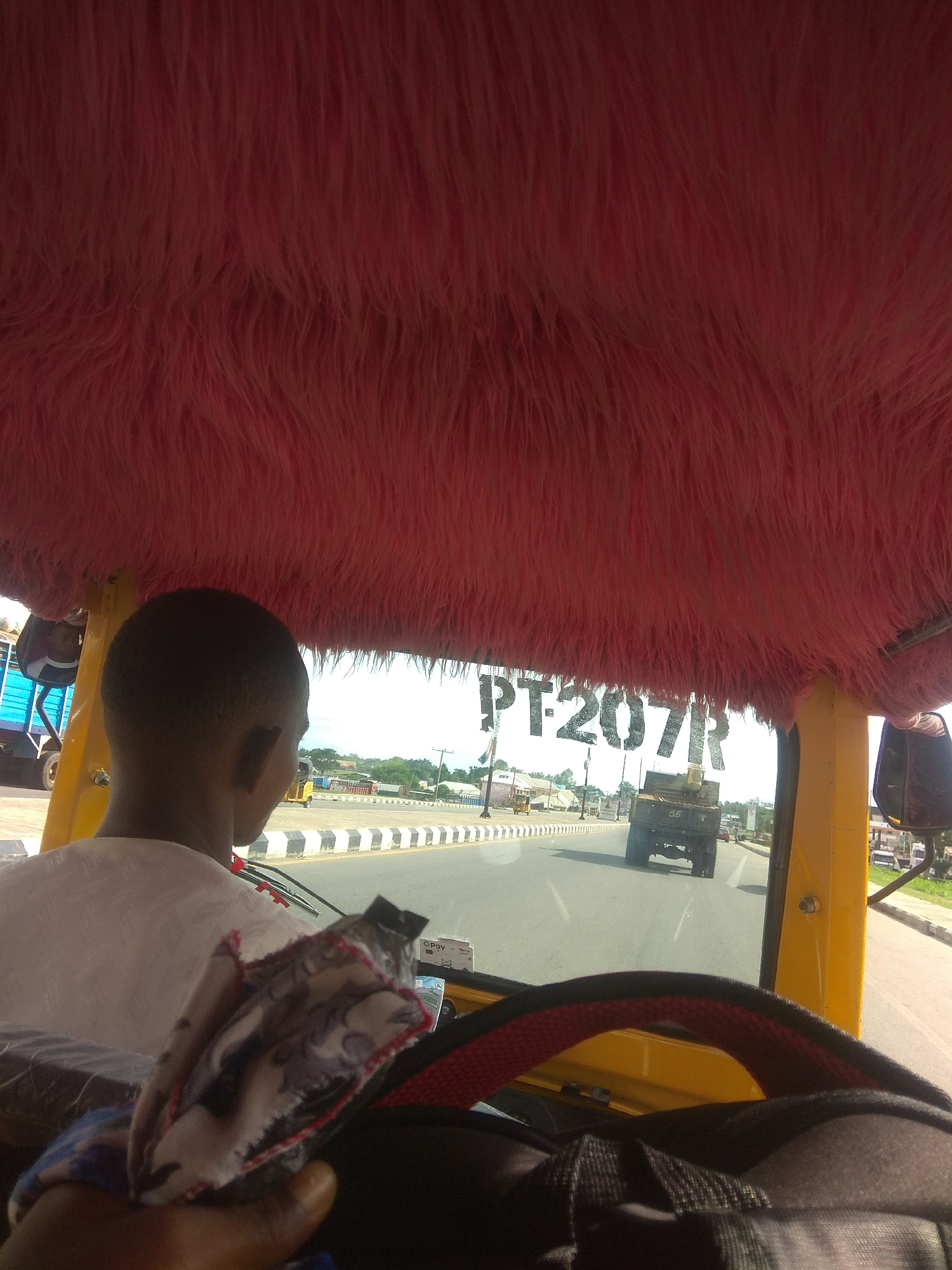 The image provides an interior view from the passenger or driver's seat of a motorized rickshaw, commonly known as an "okada" or "keke napep" in Nigeria. The ceiling of the vehicle is covered with bright pink faux fur. In the foreground, a person's head and shoulder are visible from the rear, facing towards the right. Their head is short-haired and dark. A cloth with a patterned design, possibly a handkerchief or fabric scrap, is held in the lower right portion of the frame, obscuring the steering wheel.
Through the windshield, a paved road with a median strip marked by black and white diagonal stripes is visible. Several vehicles are on the road, including a dark green truck with a cargo bed. In the background, there are buildings, trees, and utility poles. The sky is bright and overcast, suggesting it is daytime.
The license plate of a vehicle, visible through the windshield, reads "PT-207R". This suggests the location might be within Nigeria, and based on the context of the vehicle type and road infrastructure, it's plausible to be in a city like Jalingo, Nigeria, where such transport is common. However, without explicit visual cues such as street signs or landmarks definitively identifying Jalingo, this remains an inference. The overall scene depicts typical urban or semi-urban traffic in a Nigerian context.