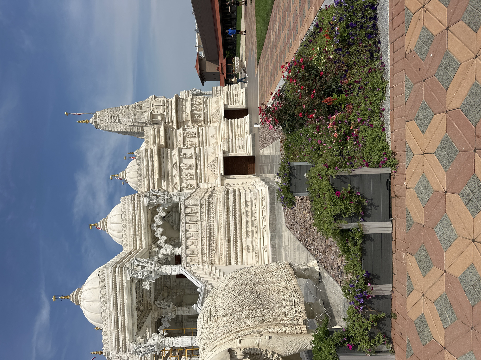 The image shows the BAPS Shri Swaminarayan Mandir in Bartlett, Illinois, USA, on a clear, sunny day with scattered clouds. The ornate white marble temple features multiple domes and intricate carvings. In the foreground, a decorative stone elephant statue is partially visible on the left, draped with a textured blanket. A planter box filled with colorful flowers, including pink, red, and purple varieties, is situated in front of the temple's entrance. The ground is paved with a patterned walkway of red and tan bricks. In the background, other buildings with red roofs are visible, and several people are walking on the grounds.