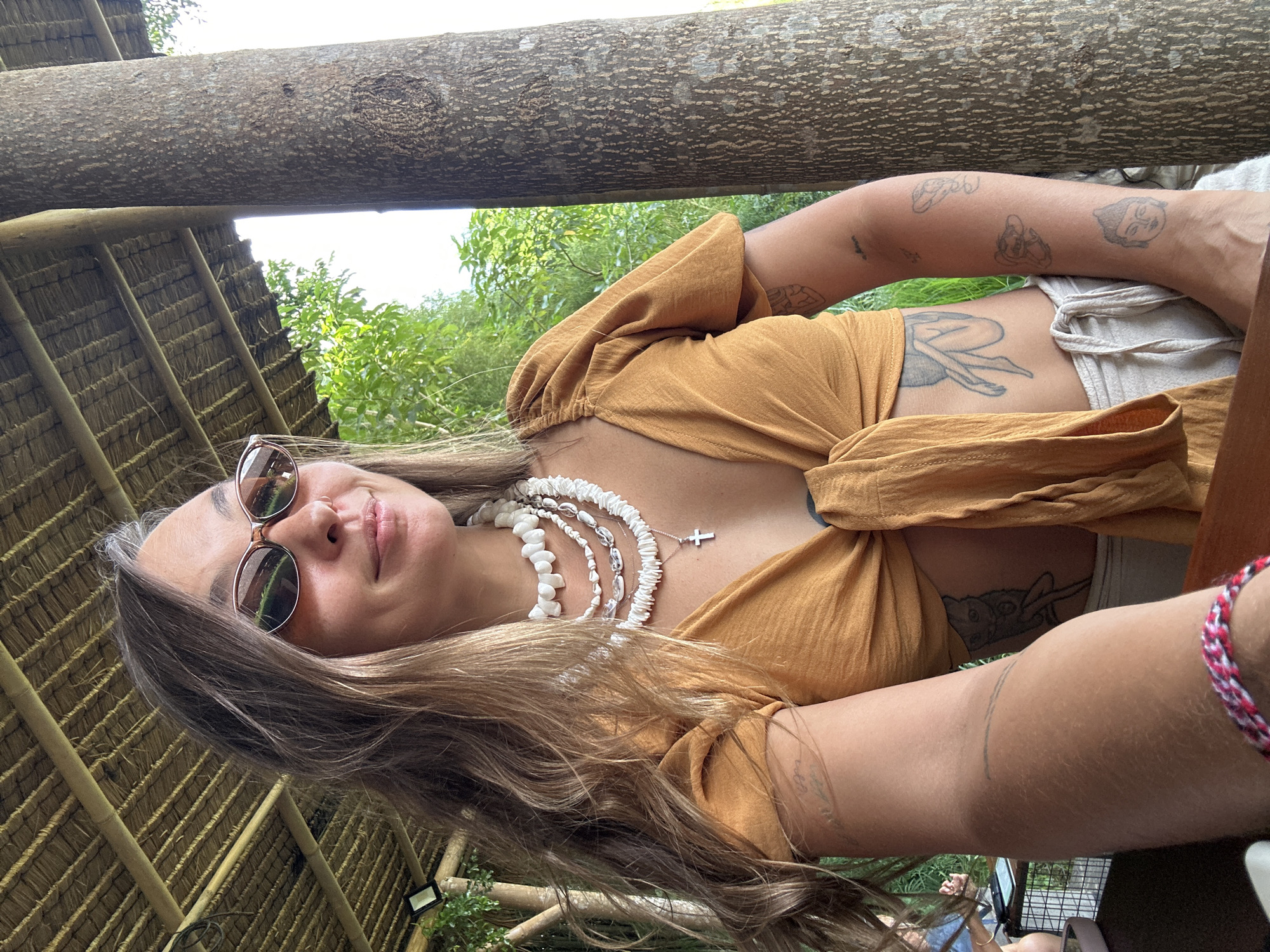 A young woman wearing sunglasses, a knotted yellow top, a white shell necklace, and a bracelet is positioned under a bamboo structure with a thatched roof. She is looking directly at the camera. The background includes lush green vegetation, possibly trees, and a wooden beam. The location is likely in the Ubud District of Indonesia, given the architectural style and the presence of tropical plants. The woman is positioned indoors. She appears to be the primary subject. Tattoos are visible on her arm and midriff.