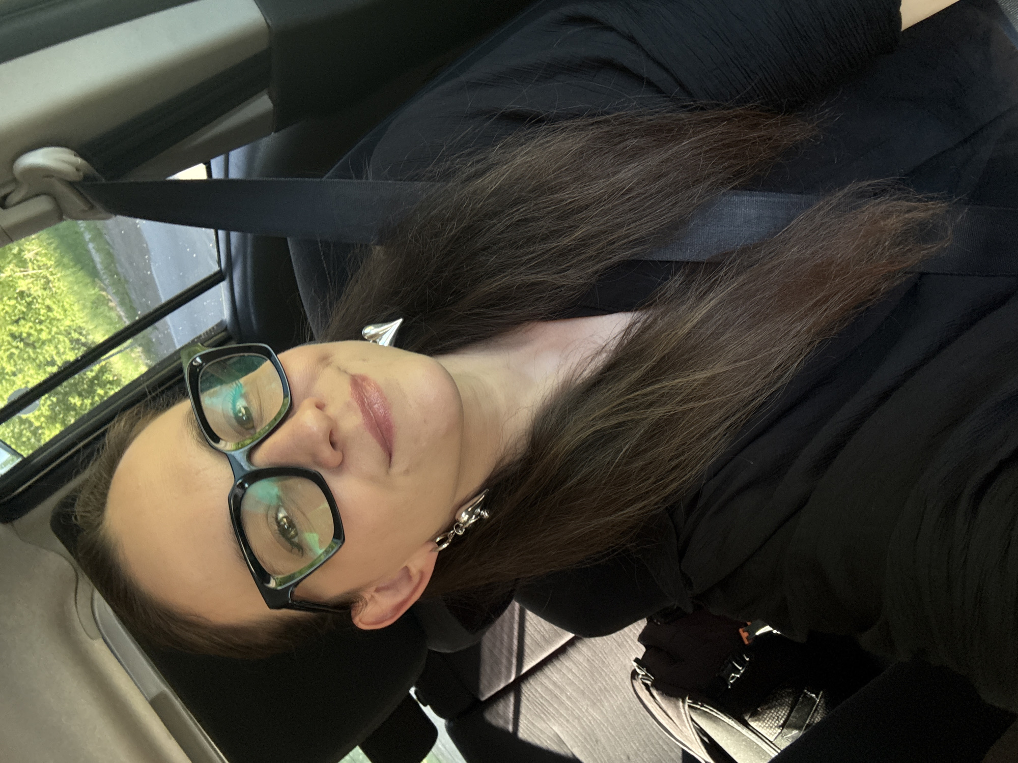 The image shows a woman wearing large black-rimmed glasses and a black top, sitting in the passenger seat of a vehicle. She has long, dark brown hair and is wearing heart-shaped earrings. A seatbelt is visible across her chest. The interior of the car appears to be dark, with sunlight streaming in from the side window, illuminating the woman's face and hair. The background outside the window shows greenery and a road, suggesting the car is in motion. The image is a selfie, taken from a slightly upward angle. There is no visible indication of the specific location in the city of Chaikovsky, Russia.