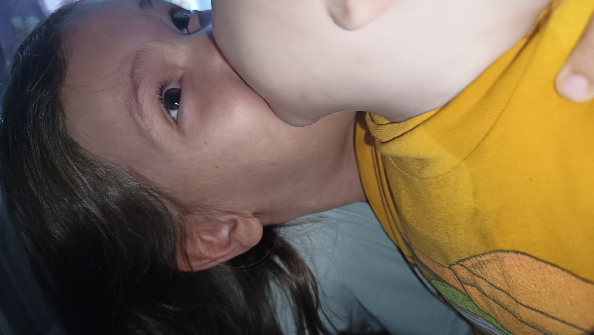 The image shows a close-up, side-view of a child breastfeeding from a person's chest. The child's face is turned away from the camera, with their eye and a portion of their cheek visible. Their dark hair is in the foreground. The person they are breastfeeding from is wearing a yellow t-shirt. The background is dark, with a hint of what appears to be a light blue sheet or blanket. The lighting suggests an indoor setting, possibly at night or in a dimly lit room. The overall tone is intimate and focuses on the act of breastfeeding. There is no visible text in the image. The location context of Karsha, Russia, is provided but not visually discernible from the image itself.
