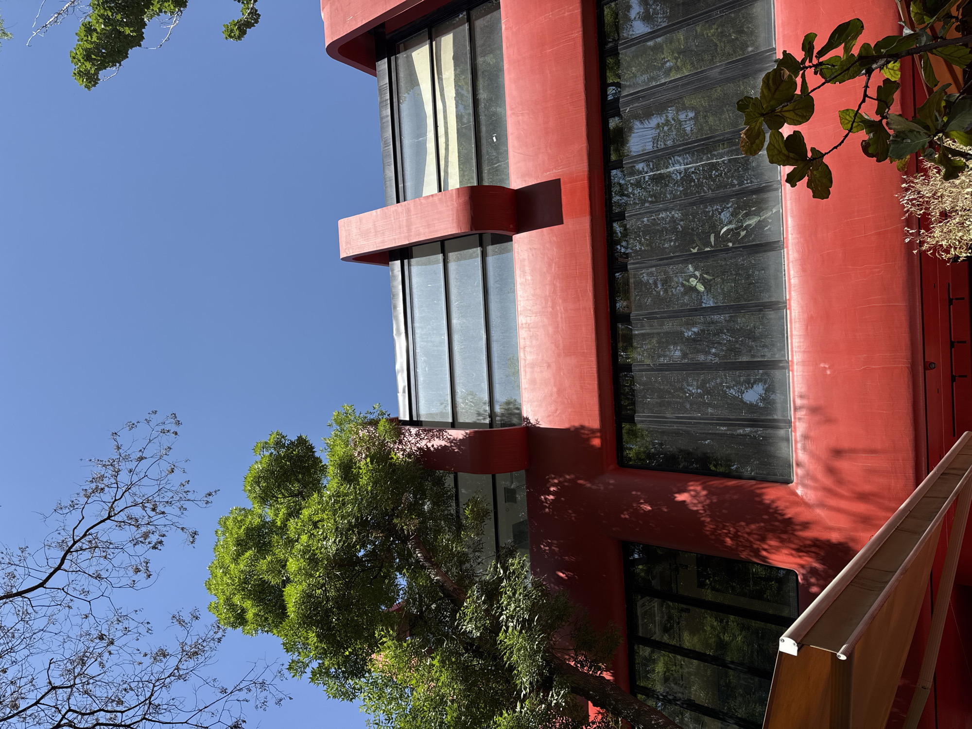 The image shows a section of a red building with large windows, surrounded by trees and set against a clear blue sky. The building has a distinct architectural style, possibly modern or post-modern, with geometric forms and a reddish-orange color. The windows reflect the surrounding trees and sky. The trees are both in full leaf and bare branches, suggesting different species or stages of foliage. The location is possibly in Yerevan, Armenia.