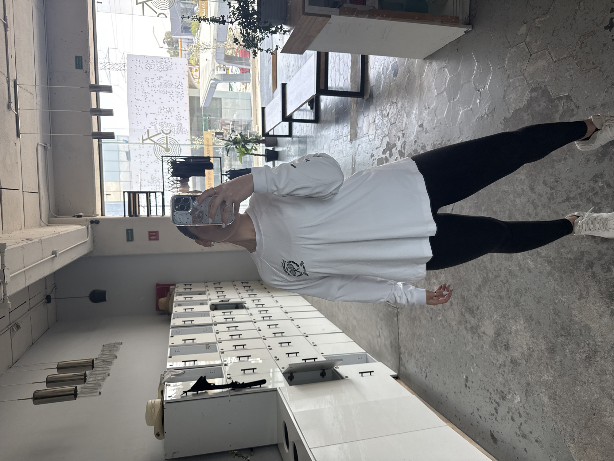 A person wearing a long-sleeved white shirt and black leggings is taking a selfie in what appears to be an indoor space with lockers and windows. The floor is a light gray concrete with a hexagonal pattern. The background shows glimpses of an urban area, potentially in Puebla, Mexico, due to the architecture and vegetation seen through the windows. The selfie is taken indoors. There are lockers lining the left side of the scene. The person seems to be alone in this indoor space.