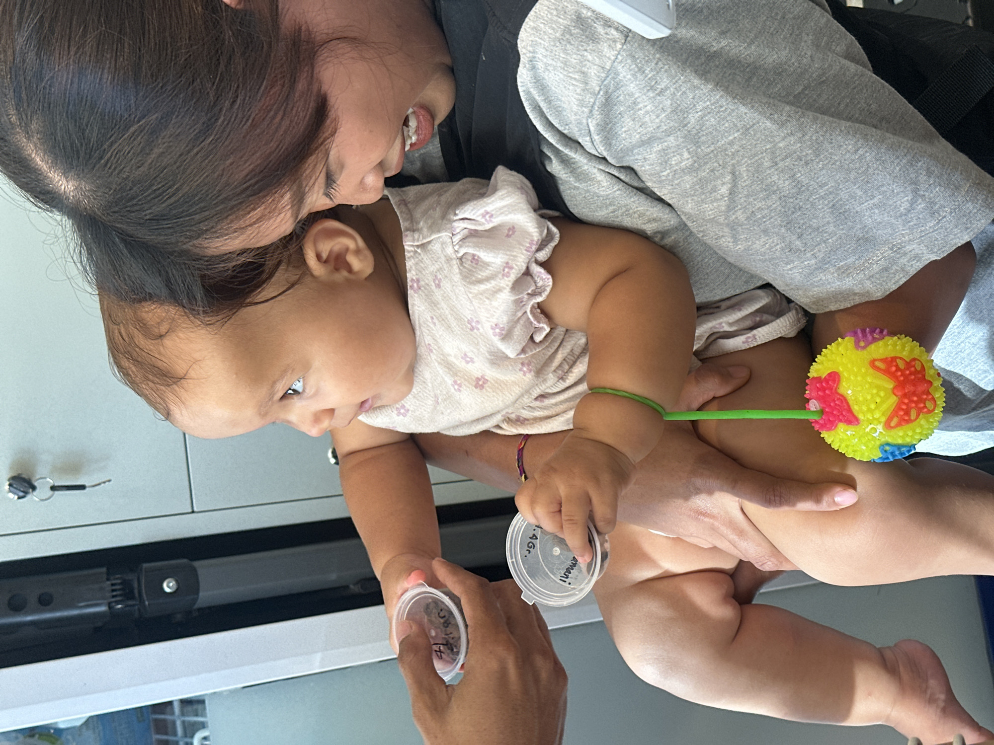 The image shows a baby being held by an adult. The baby is wearing a light-colored floral patterned shirt. Another adult is seen in the background, partially obscured. A colorful spiky ball toy is visible near the baby's legs. Two small, clear plastic containers with handwritten labels and contents, possibly organic material, are being held near the baby. The location is North Kuta, Indonesia, based on information associated with the image. The scene suggests an interaction involving an adult holding the baby, while another adult handles containers with samples.