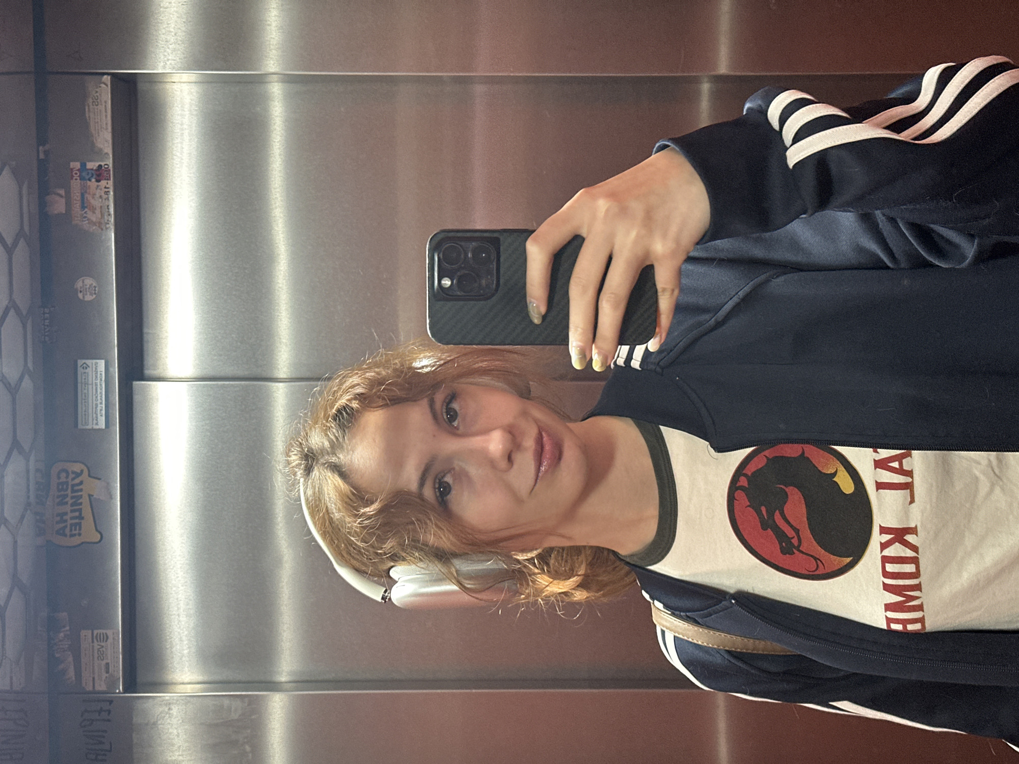 A woman is taking a selfie in an elevator. She is wearing a navy blue Adidas tracksuit jacket with white stripes on the sleeves over a white t-shirt featuring a Mortal Kombat logo. She is holding a black smartphone with a multiple-lens camera. She has headphones around her neck. The elevator interior is stainless steel with mirrored sections. The woman appears to be in her late 20s to early 30s with brown hair. Labels and stickers are visible on the elevator door. The location is indicated by the presence of Novi Sad, Serbia, specific advertisements.