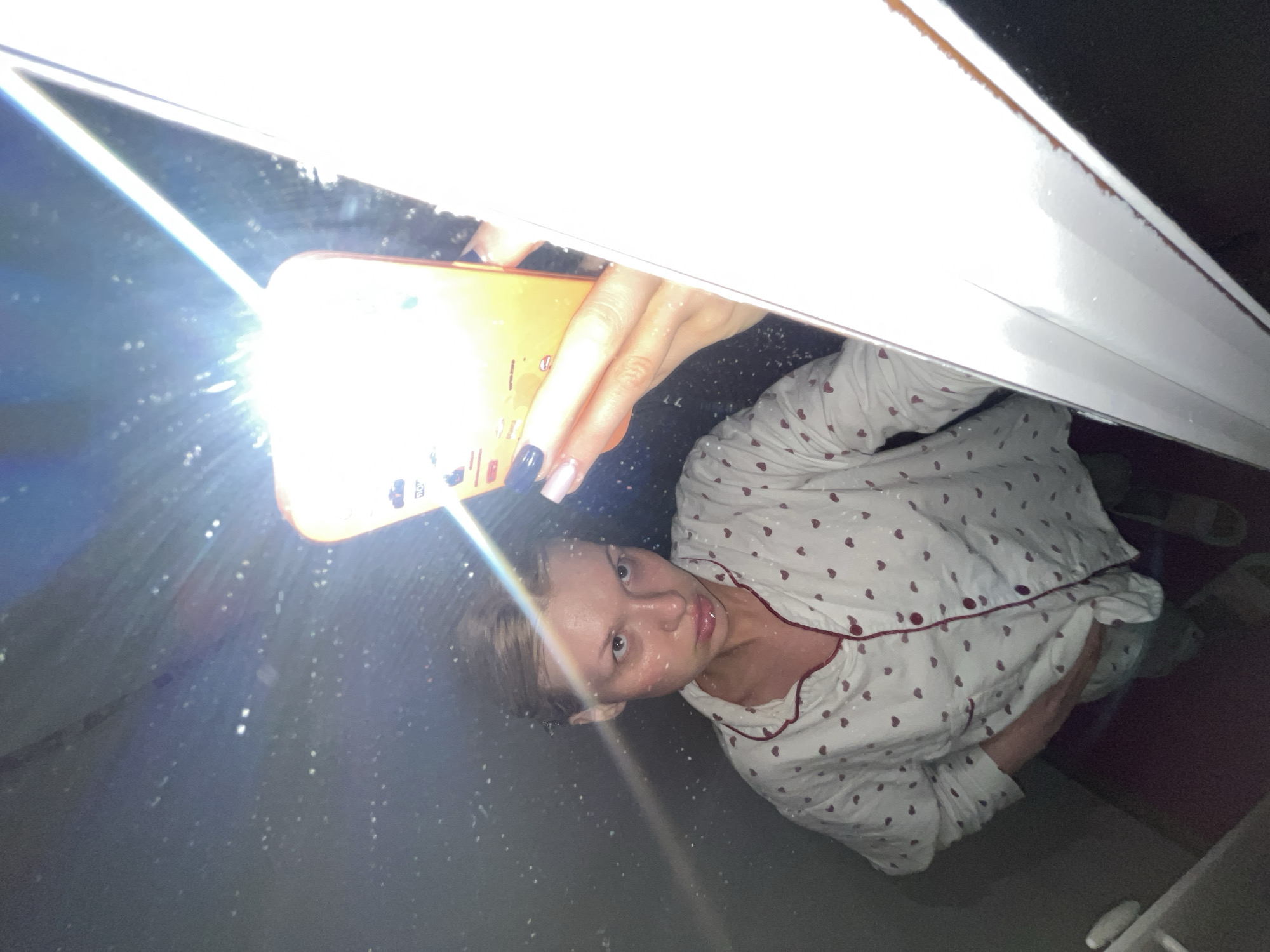 A woman wearing white pajamas with red hearts is illuminated by a bright smartphone flashlight. She appears to be positioned below a window frame or some other horizontal structure. The location is in the city of Chaikovsky, Russia.