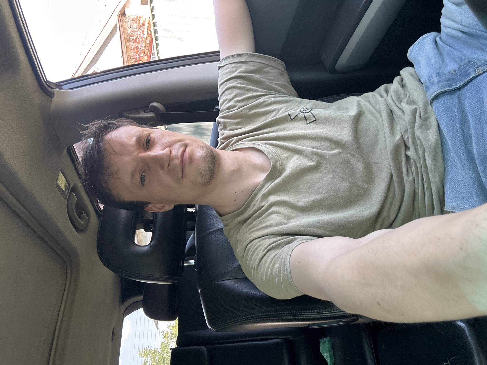 A Caucasian male with light brown hair and stubble is taking a selfie from the driver's seat of a car. He is wearing a light olive green t-shirt with a black graphic on the left chest and blue jeans. The car interior is visible, with black leather seats, a sunroof, and part of the passenger side door and window. Outside the window, a building with a red tiled roof and a white fence can be seen, consistent with a residential area in Makiv, Ukraine. The lighting suggests it is daytime.