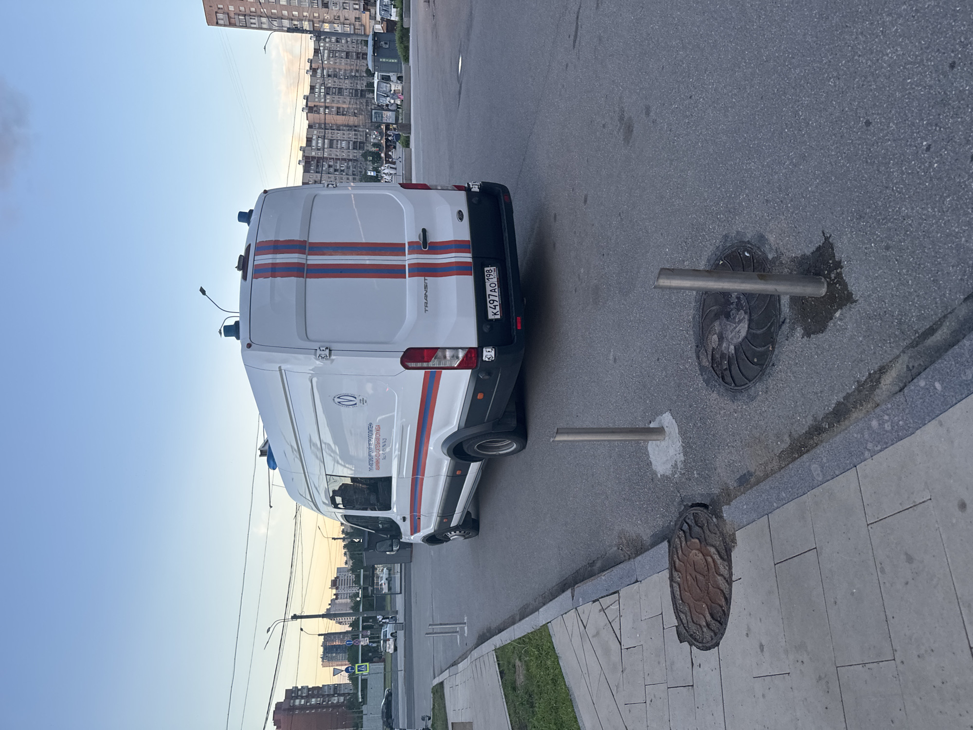 A white Ford Transit van with red and blue stripes on the rear doors is parked on a paved surface in Saint Petersburg, Russia. The van has "TRANSIT" written on the rear and a Russian license plate K497AO 198. On the side of the van, text reads "ГУП «ПЕТЕРБУРГСКИЙ МЕТРОПОЛИТЕН» АВАРИЙНО-ДИСПЕТЧЕРСКАЯ СЛУЖБА". There are two blue lights on the roof of the van. The van is parked next to a sidewalk with a manhole cover and a metal bollard. In the background, there are apartment buildings and several other white vans or minibuses. The sky is a pale blue with some clouds, suggesting it is either early morning or late evening. The scene appears to be a street or parking lot in an urban environment.