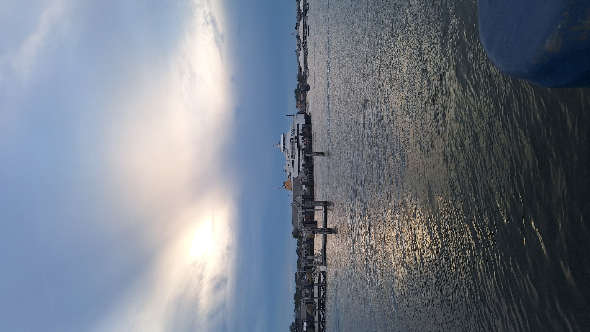 A ferry is docked at a harbor on Nantucket Island, Massachusetts. The sky is overcast with a bright patch of sunlight peeking through the clouds. Several boats are visible in the background, and the water is choppy with reflections of the sky. The ferry is white with a yellow funnel and is positioned on the right side of the image. Buildings and docks are visible along the shoreline.