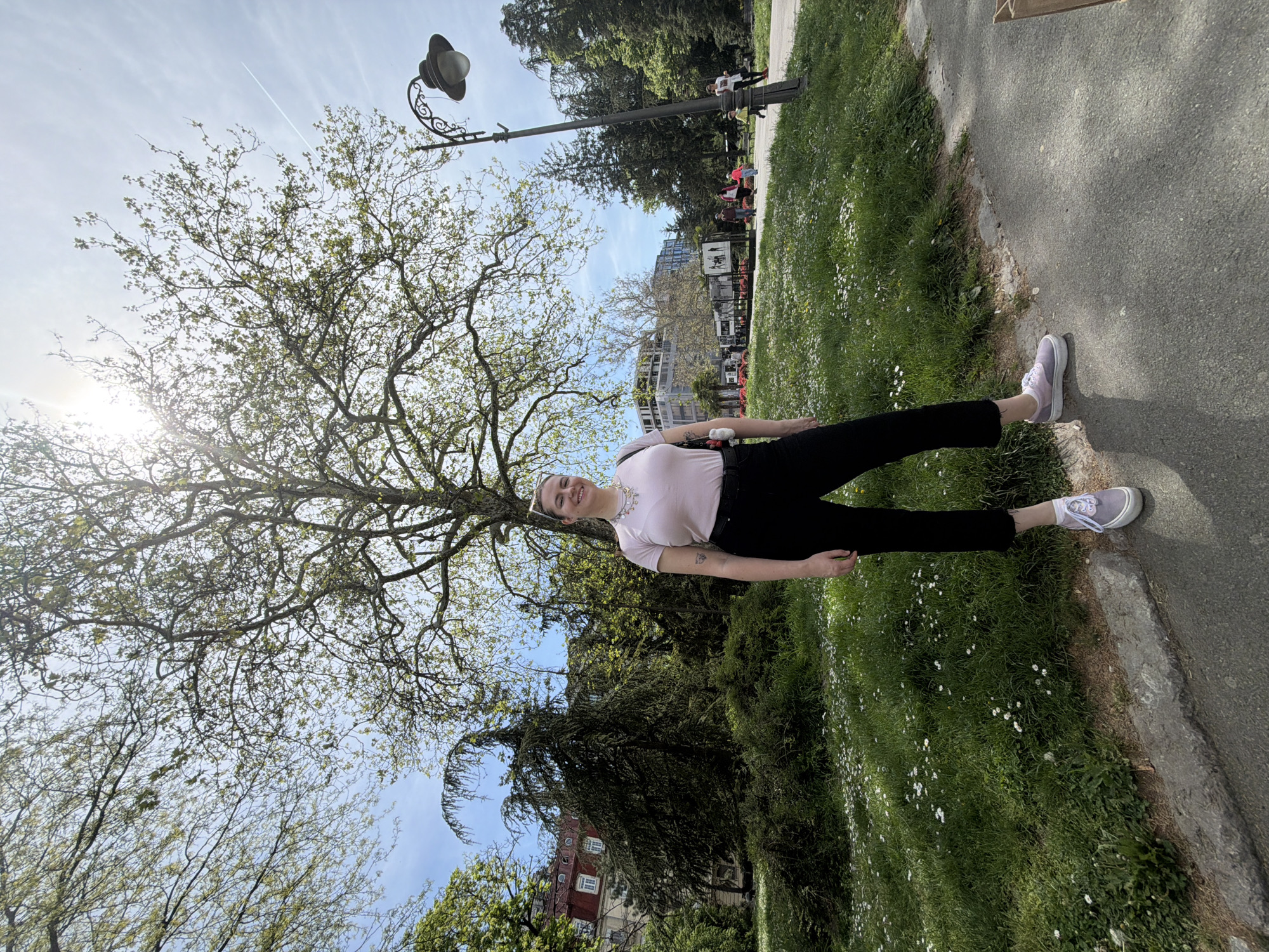 A young woman stands in a park in Belgrade, Serbia, on a sunny day. She wears a light pink t-shirt and black pants, with gray tennis shoes. A tree with budding green leaves dominates the left side of the frame. In the background, other trees and a light post are visible, along with a building with red roofing. The ground is a mix of grass and a gray paved path. Small white flowers dot the grassy areas. People can be seen in the background further down the path. The sun is shining brightly, creating a lens flare effect on the left side of the image.