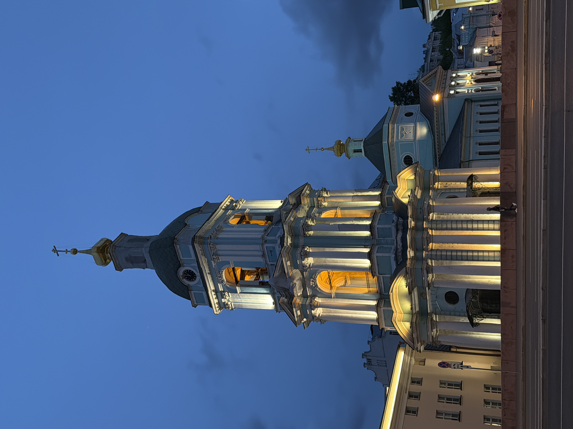 The image showcases a tall, ornate church tower against a twilight sky. The tower has a distinctive blue and white facade, with multiple levels featuring columns and arched openings illuminated by warm light. A green dome sits atop the tower, capped with a golden spire and a cross. To the right of the main tower, a smaller, similarly styled structure is visible. In the background, city buildings line the street, illuminated by streetlights. The scene is set in Перовское сельское поселение, Ukraine.