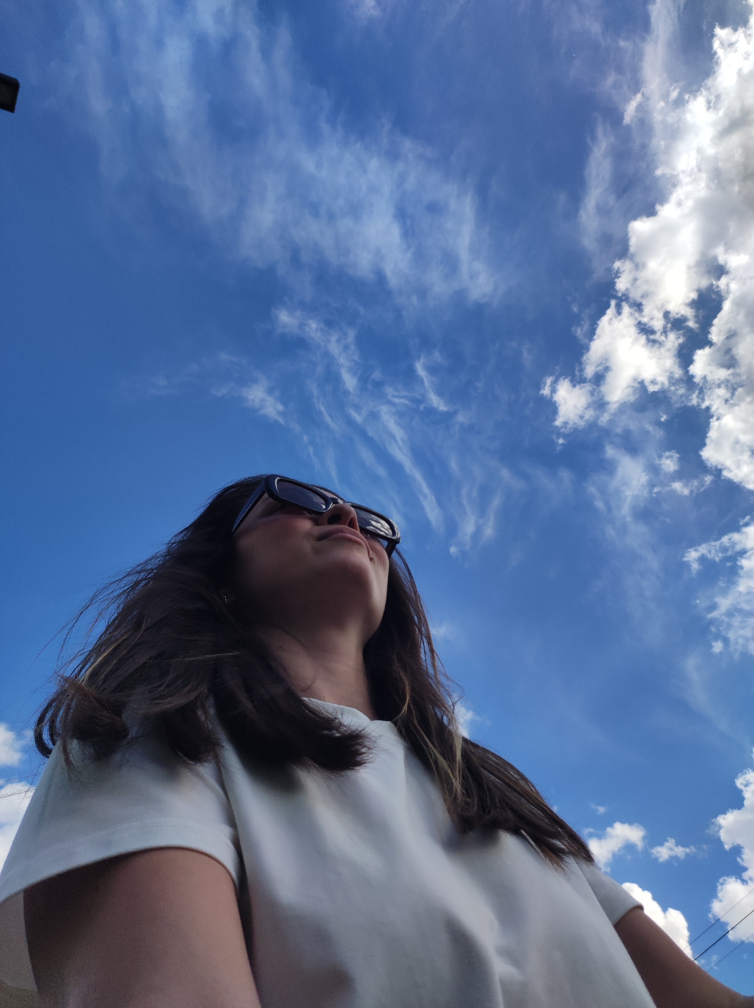 A low-angle photograph shows a person looking up at a partly cloudy blue sky. The person has dark hair with lighter streaks, is wearing dark sunglasses, and a white t-shirt. The sky above is blue with scattered white clouds, with some cirrus formations. A pole or mast is visible in the upper left corner. Based on the visual information, the photograph location within the city of Chaikovsky, Russia cannot be determined.