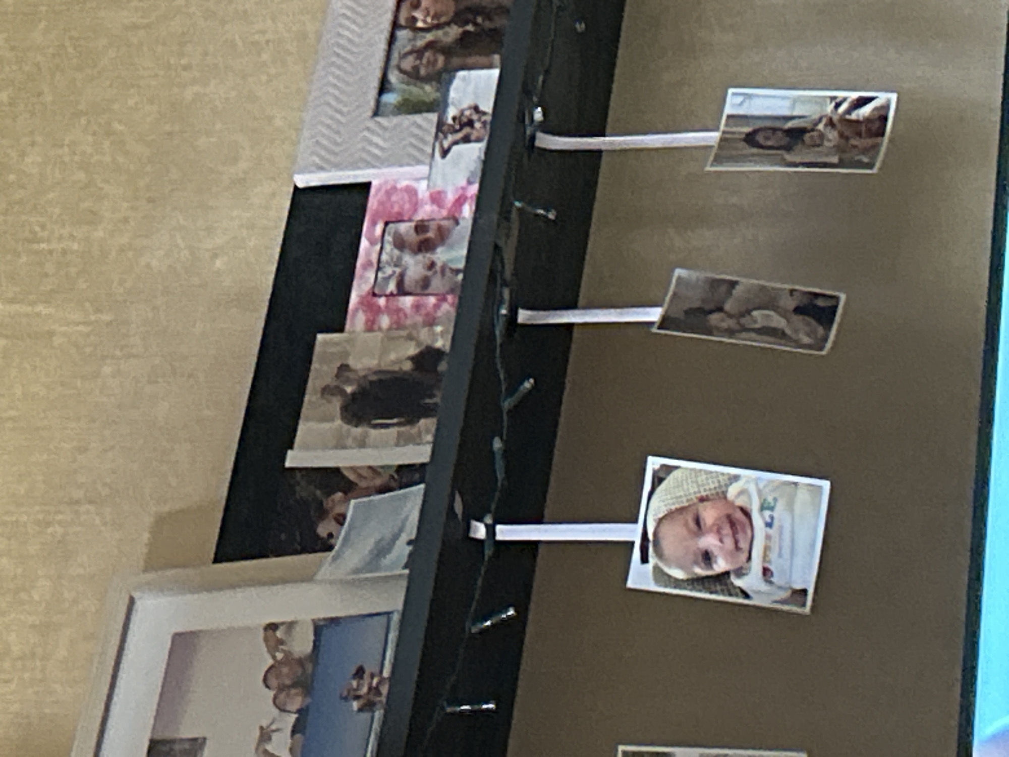 This media file depicts a collection of photographs displayed on a dark surface, possibly a wall or a frame. The photographs are of various sizes and orientations, and some appear to be held up by white strips of paper, which are attached to what look like fairy lights.
The subjects in the photographs are predominantly people, with a notable focus on a baby in one of the most prominent images. This baby is wearing a light-colored hat and clothing with some writing on it. Other photos show groups of people, including what appear to be adults and children, in different settings. One image shows a person lying down, possibly in a crib or bed. Another photo displays a close-up of a person's face. In the lower left, a framed photograph is partially visible, showing several people with their arms around each other, suggesting a celebratory or family gathering.
The overall impression is one of personal memories and family life being showcased. The fairy lights add a decorative and festive touch. The background is a textured, light-colored surface that contrasts with the darker display area and the photographs themselves. There is no indication of the time of day, weather, or specific outdoor location. However, the context provided (Sofia, Bulgaria) suggests the photos are likely from this region. The emotion conveyed is generally positive, hinting at happy memories and familial bonds. No significant text is clearly legible in the visible photographs, other than potentially on the baby's clothing.
