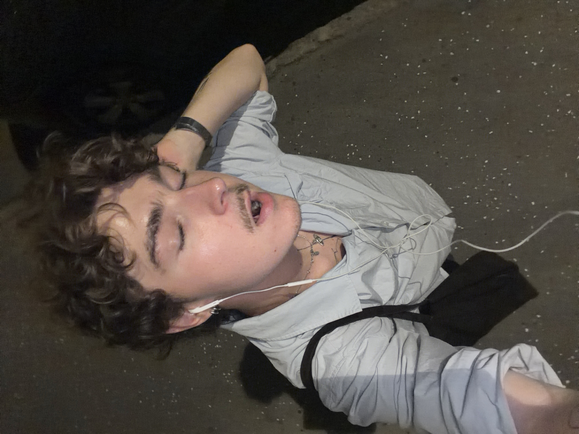 A young person with curly brown hair and closed eyes is shown from a high-angle perspective, outdoors at night. They are wearing a light blue collared shirt and white earbuds. The person has a mustache and appears to be singing or speaking with their mouth open, with their right hand touching their head. A dark bag is slung over their left shoulder. The background consists of a dark, asphalt-like surface with scattered white debris and a hint of a dark object, possibly a car, in the upper right corner. The ground appears to be slightly elevated on the right side, suggesting a sidewalk or curb. The lighting is dim, with the person's face and upper body illuminated. The presence of asphalt, a sidewalk, and a car suggest an urban setting within Vracar Urban Municipality, Serbia.