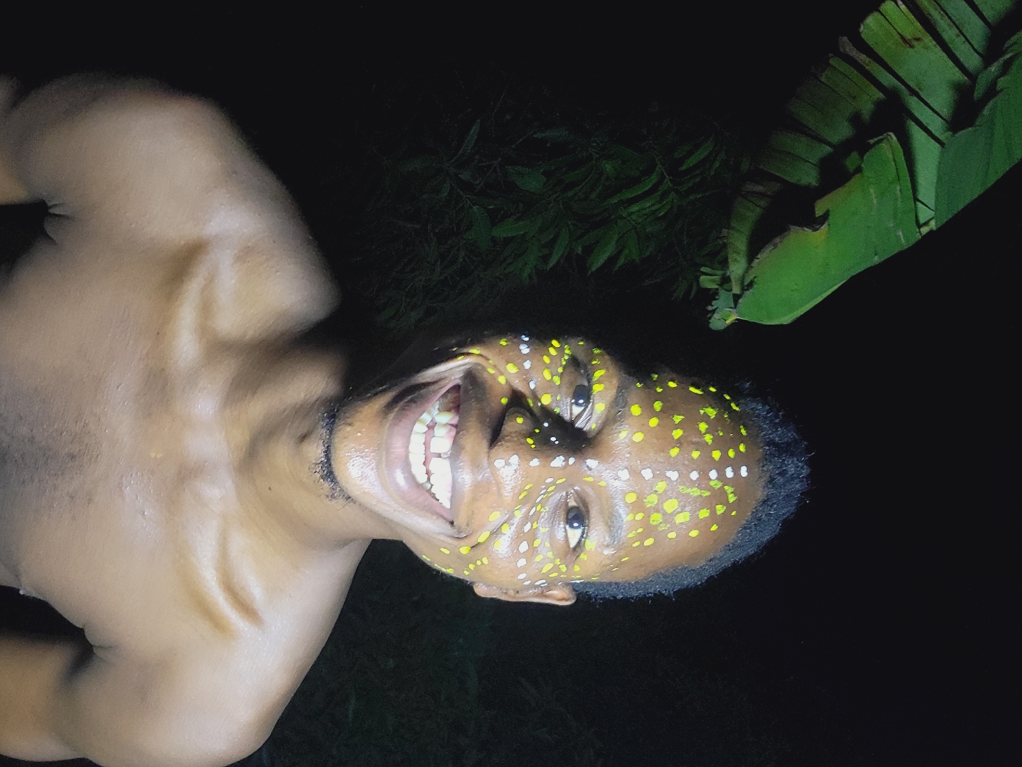 An eye-level, rotated shot depicts a shirtless dark-skinned man with white and yellow dots painted on his face. The subject is smiling, revealing his teeth. To the left is his exposed shoulder and neck. Behind him, there are leafy plants including what appears to be a banana leaf. The location is unspecified but assumed to be in Nkpor, Nigeria. The overall lighting is dim, suggesting a nighttime setting.