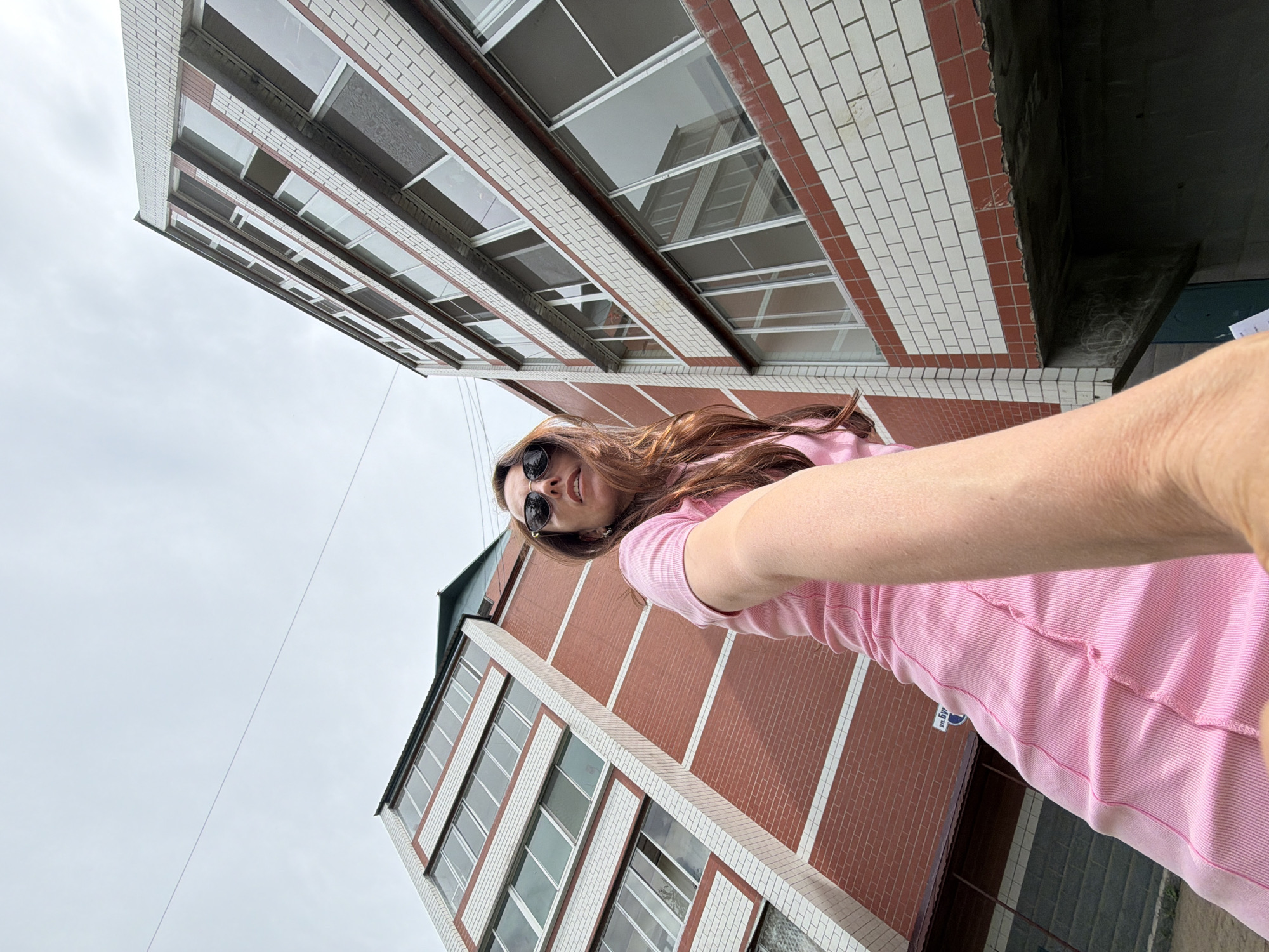 In the city of Birobidzhan, Russia, a woman wearing sunglasses and a pink t-shirt is photographed from a low angle, looking up at the camera. She stands outdoors in front of a modern apartment building with a facade of red and white brick. The building features multiple floors of windows, some of which reflect the sky and surrounding structures. The sky above is overcast. The woman's right arm is extended towards the camera, creating a selfie-like perspective. The overall scene suggests an urban environment with a focus on architectural details and the individual.
