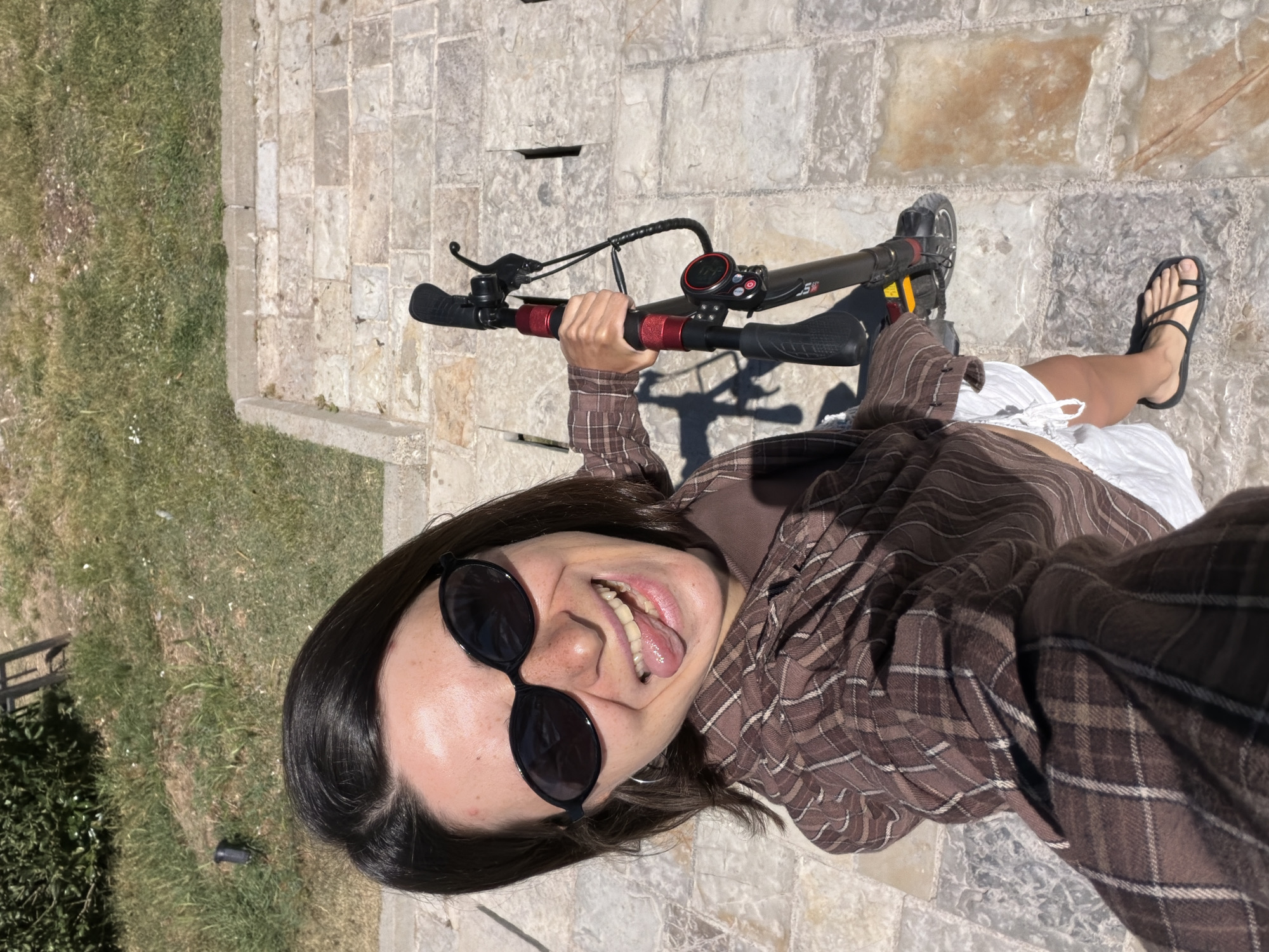 A ground-level, high-angle medium shot shows a woman posing for a selfie in what may be the city of Budva, Montenegro. She's facing the camera and grinning, sticking her tongue out. She wears dark sunglasses, a brown plaid shirt over a brown top, and white shorts. Black strappy sandals are visible on her feet. She is standing on a stone tile surface. In her hands, she holds the handlebars of an electric scooter. The scooter is black and red, with a digital display. To her left is a patch of green grass. The environment appears to be outdoors on a sunny day.