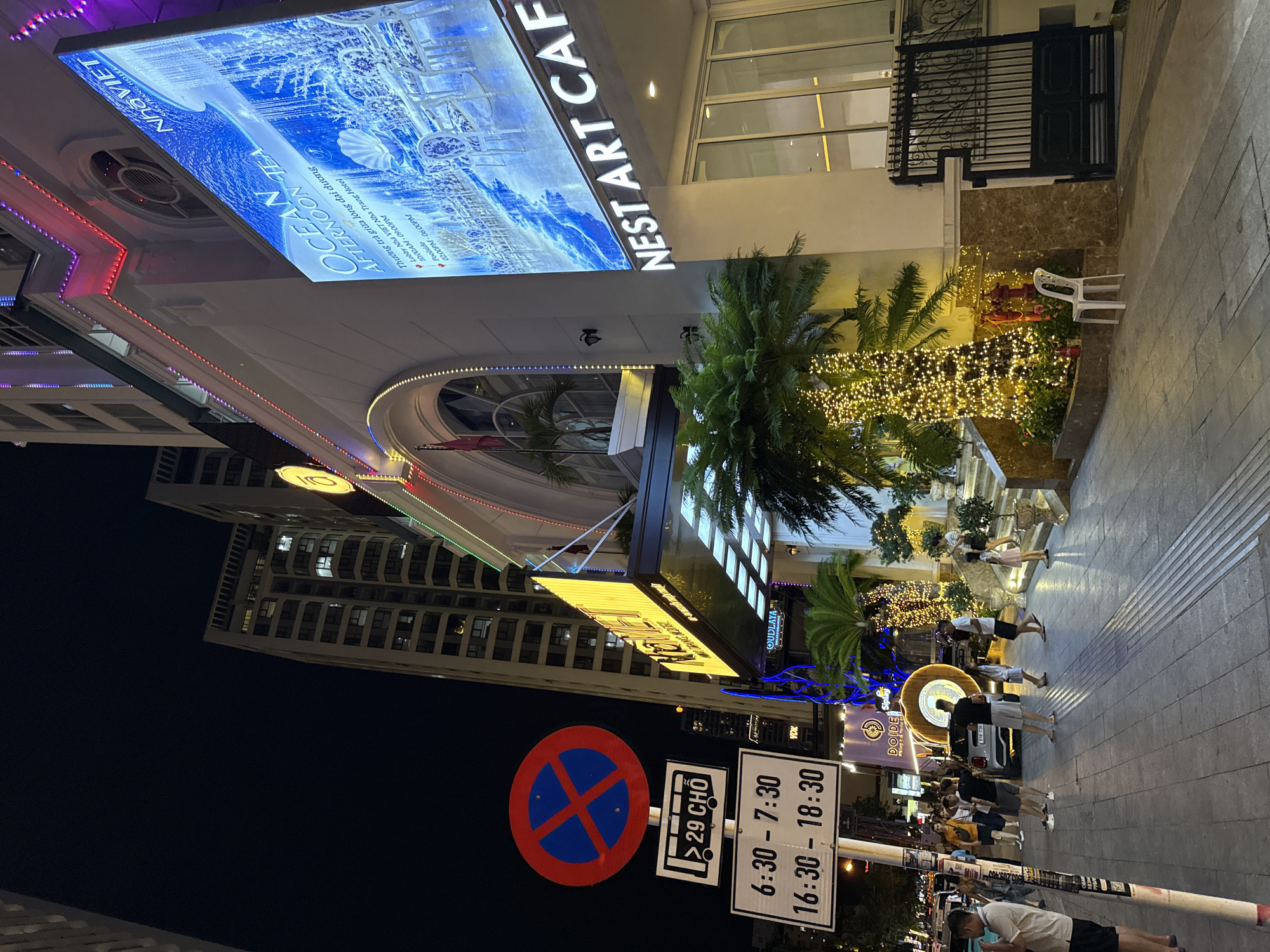 The image depicts a nighttime scene on a street in Nha Trang, Vietnam, likely in front of a hotel or establishment called "NEST ART CAFE". A prominent, brightly lit advertisement for "OCEAN AFTERNOON TEA" at "NHA VIET" is displayed on the right side of the building. The building itself is decorated with colorful lights, particularly along its facade. On the left side of the street, there are traffic signs, including a no-parking sign and signs indicating bus capacity ("29 CHO") and parking restrictions with specific times: "6:30 - 7:30" and "16:30 - 18:30". Several people are visible walking on the sidewalk, some in casual attire, and others appearing to be patrons of the establishment. A small car with illuminated features is parked further down the street. Palm trees, some adorned with string lights, are present along the sidewalk and in front of the building, contributing to the festive atmosphere. The street appears to be paved, and there are streetlights casting shadows. The overall environment suggests a busy urban area at night, with commercial activity and pedestrian traffic.