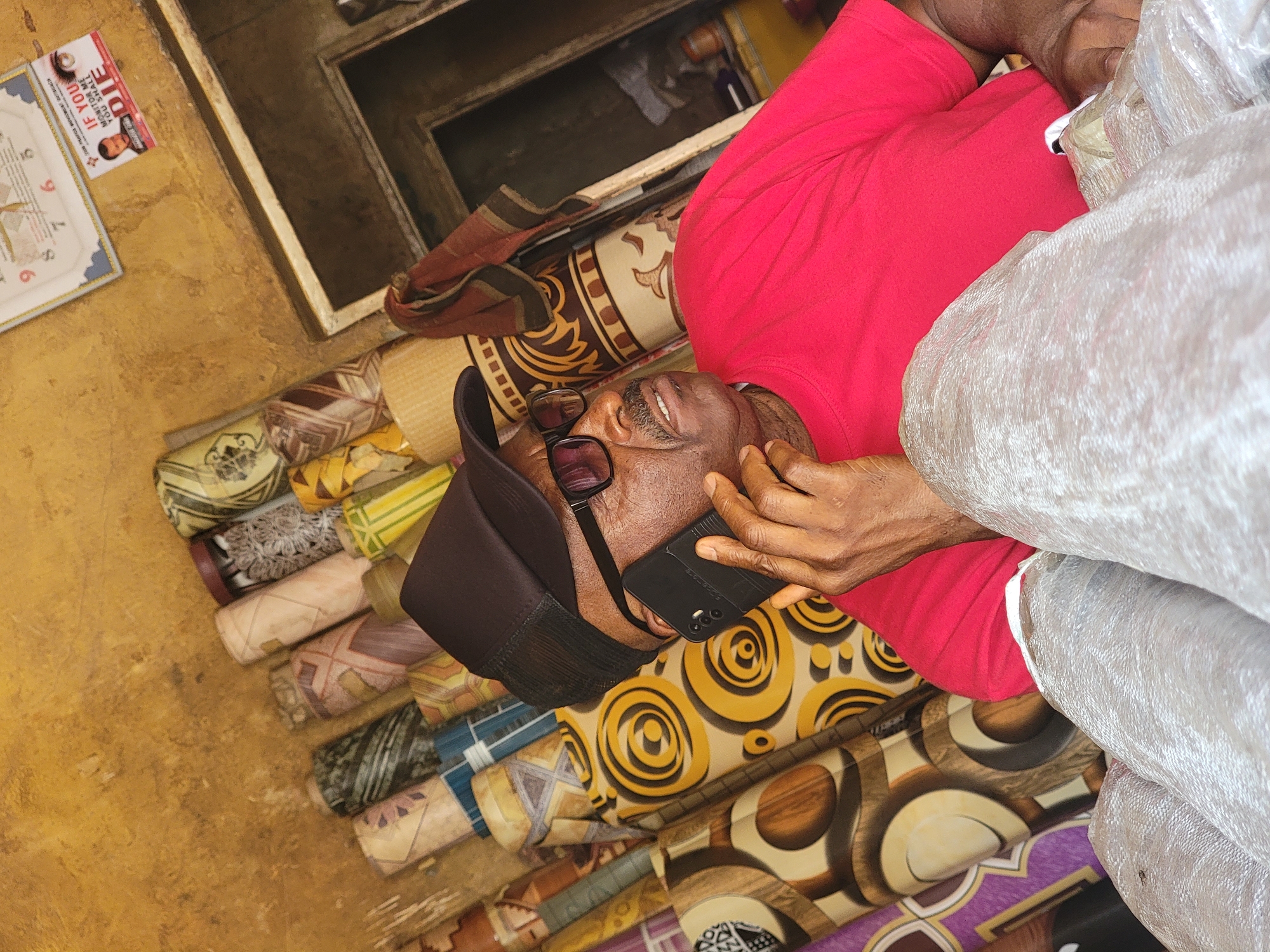 An eye-level indoor shot of a man with dark skin, wearing a red shirt, black glasses and a black cap is on the phone in Maiburiji, Nigeria. The man is surrounded by rolled-up carpets of different patterns and colors. The man is holding a black smartphone to his right ear. Some of the carpets have yellow and brown circular patterns. There is a poster on the yellow wall in the background, as well as a window. The lighting is indoor.