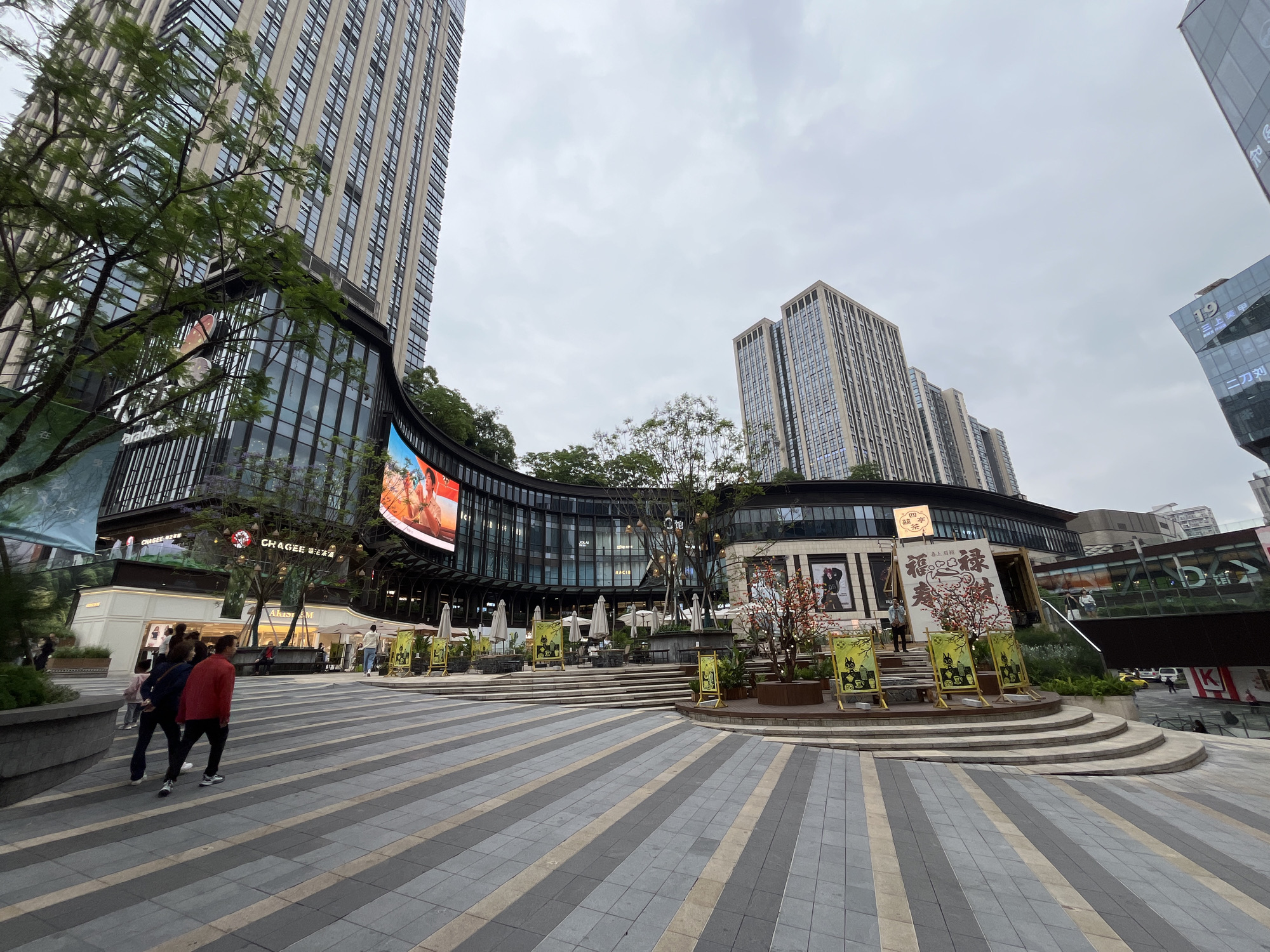 The image shows an outdoor urban space with tall buildings and various commercial establishments. It appears to be a public square or a pedestrian zone in a city, possibly located in the Yuzhong District of China. The pavement is paved with gray and beige tiles. Two people are walking in the bottom left corner of the image. There is signage in Chinese characters and advertisements on the buildings. There are multiple restaurants with outdoor seating. The sky is overcast, suggesting that the weather is cloudy. A large LED screen is visible on the building to the left. There are landscaped areas with trees and shrubs. There is signage for the restaurant "CHAGEE." The establishments "Paradiso," "Akust," "三上美甲", and "二刀刘" are also labeled on various signs.