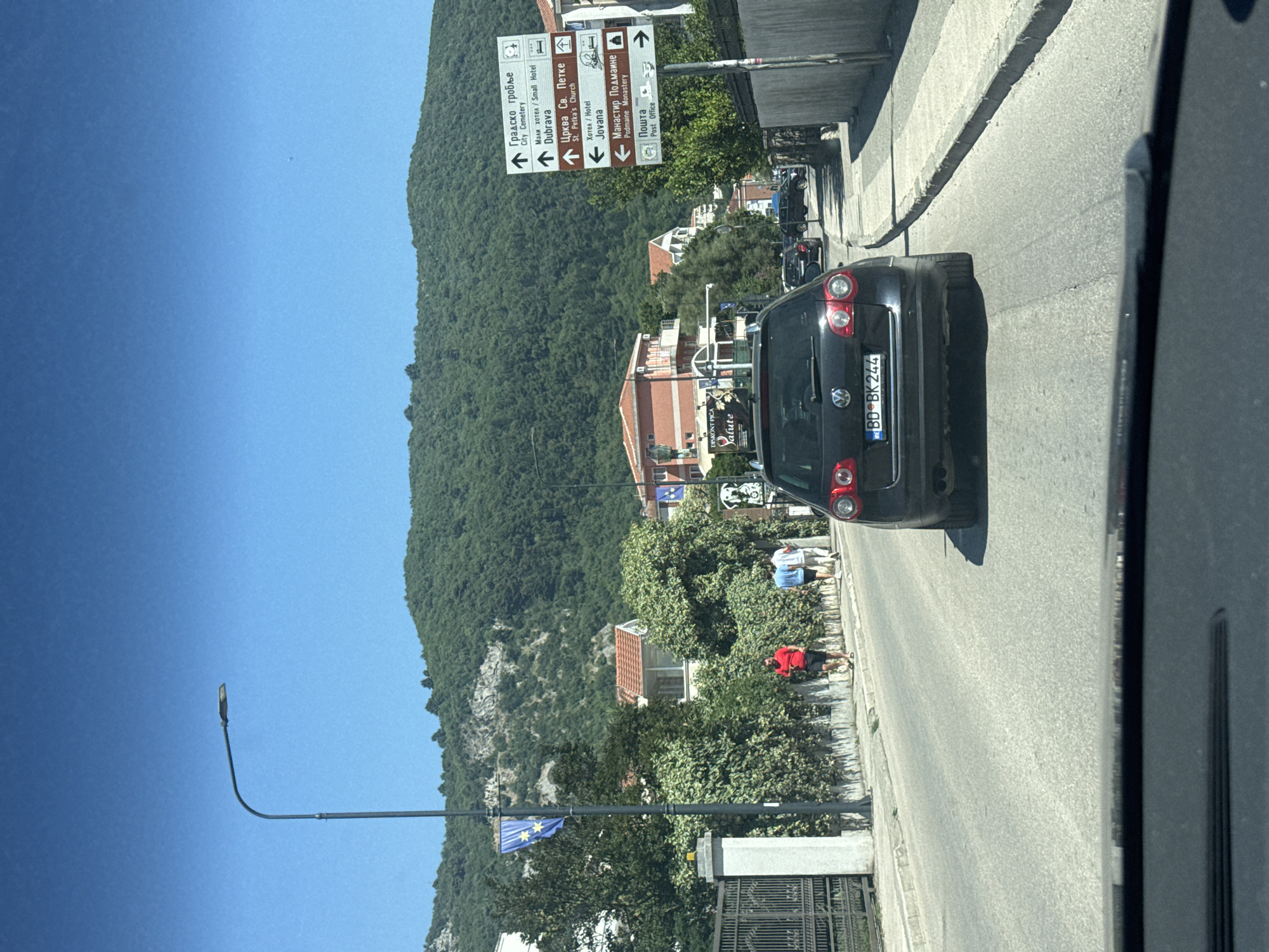 The image depicts a view from inside a vehicle traveling on a street in Budva, Montenegro. The street is paved and appears to be in good condition. On the right side of the street, there is a sidewalk with a low stone wall and then a building. On the left side of the street, there is a sidewalk with a hedge, a fence, and then buildings. The street is lined with trees and vegetation.
In the foreground, the dashboard of the vehicle is visible. A black Volkswagen Passat is in front of the vehicle, with its license plate "BD BK 244" clearly visible. The car is traveling on the road.
Several people are walking on the sidewalk on the left side of the street. One person is wearing a red shirt and black shorts, another is wearing a white t-shirt and light-colored shorts, and a third is wearing a light blue t-shirt and dark shorts.
There are several signs on the right side of the street. One signpost has multiple directional signs pointing to various locations, including "Gradsko groblje" (City Cemetery), "Mali hotel / Small Hotel Dubrava", "Crkva Sv. Petke / St. Petka's Church", "Hotel Jovana", "Manastir Podmaine / Podmaine Monastery", and "Пошта / Post Office".
In the background, there are buildings with red-tiled roofs and a lush green hill. A street lamp is visible on the left side of the road, and a flag with stars on a blue background is hanging from it. The sky is clear and blue, indicating a sunny day.
The overall impression is that of a typical street scene in a European city on a clear day. The presence of directional signs in Cyrillic and Latin scripts suggests a bilingual environment, which is characteristic of Montenegro.
